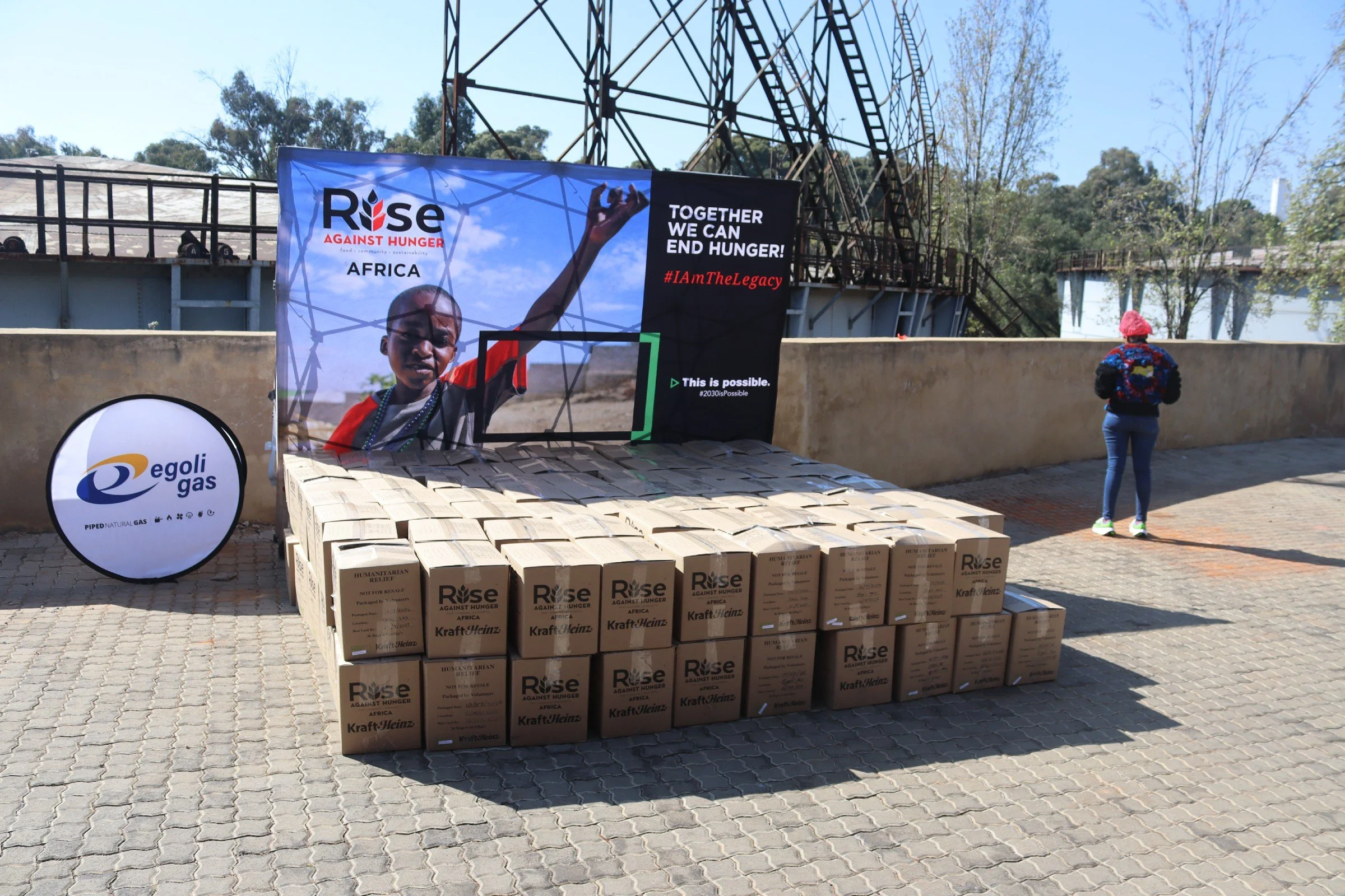 A table with many boxes of humanitarian relief supplies labeled 'Rise Against Hunger.' Behind the table, a large banner displays a young boy raising his hand with the slogan 'Together We Can End Hunger!' and hashtags #IAmTheLegacy and #2030isPossible