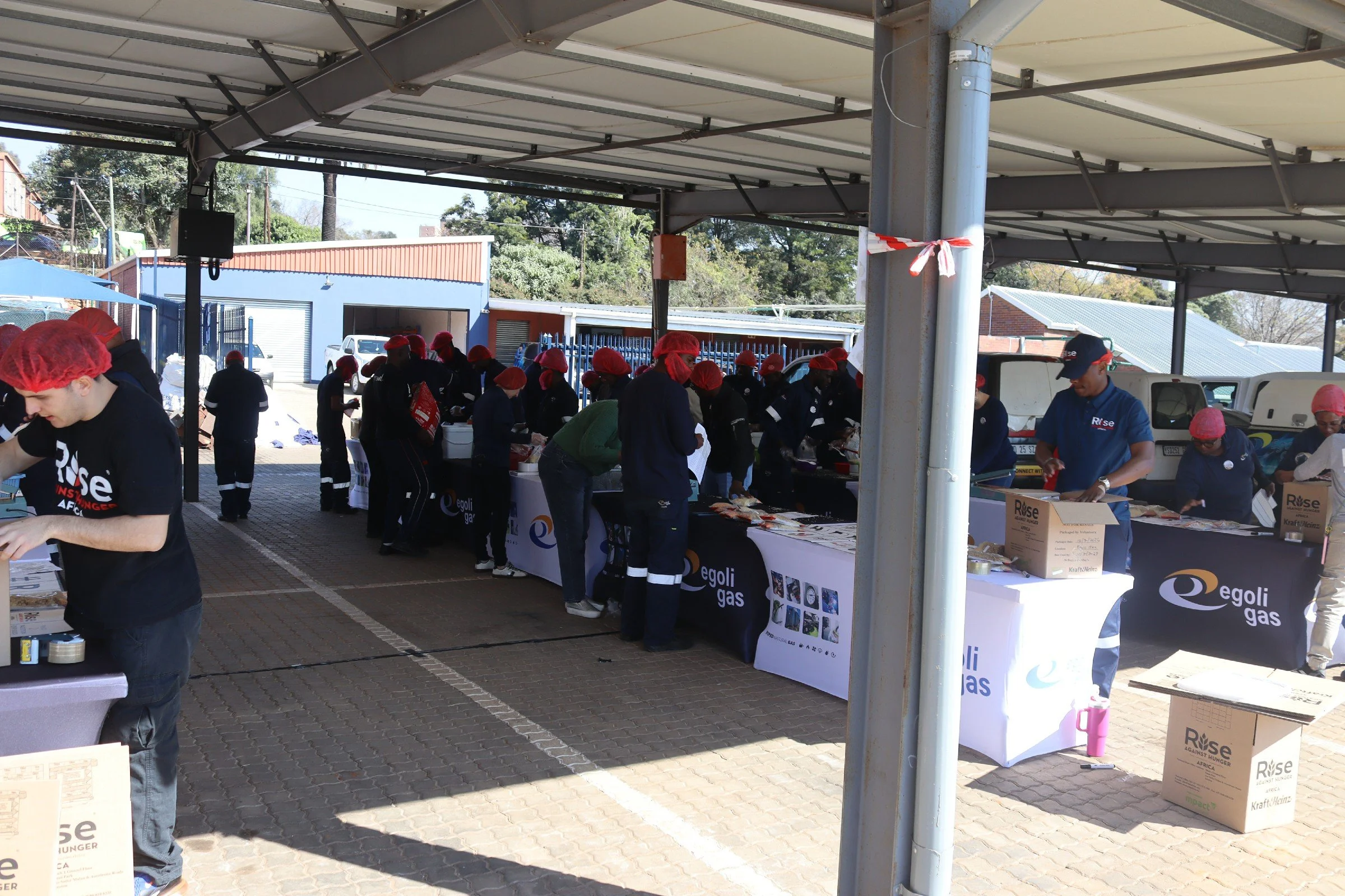 Community event with volunteers wearing red hair nets, working at tables under a shelter. Boxes and banners from Rise Against Hunger are visible, indicating a packaging or distribution activity.