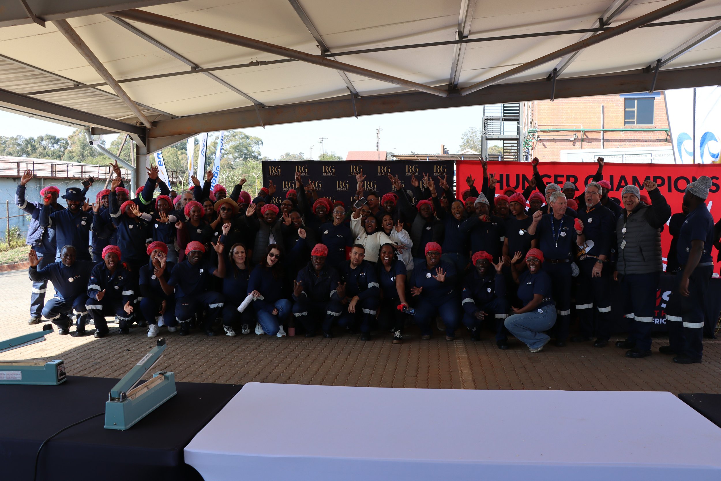 A large group of people, many wearing matching uniforms and red hairnets, are standing outside under a canopy, celebrating with their hands raised. There are banners and a sign that reads "HUNGER CHAMPIONS" in the background.