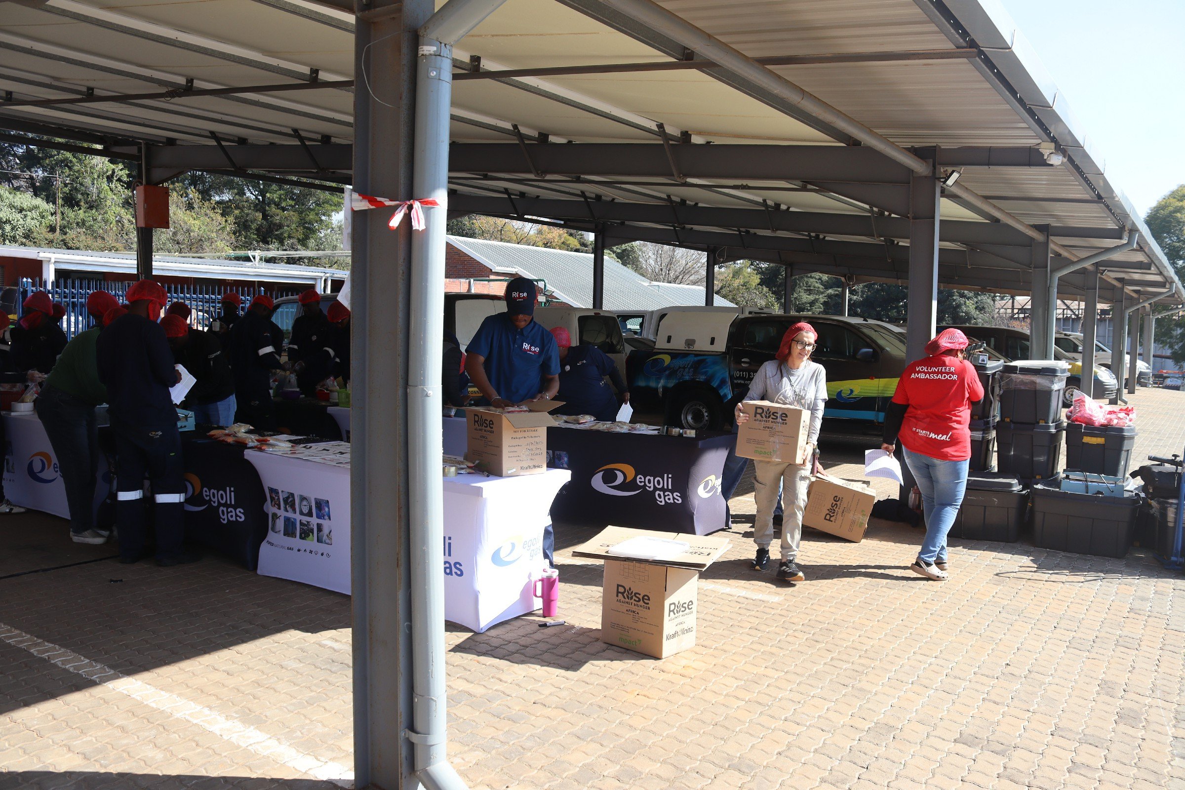 People wearing red hair caps and volunteer shirts inside a covered outdoor area with tables, boxes, and vehicles, participating in a community service or donation event.