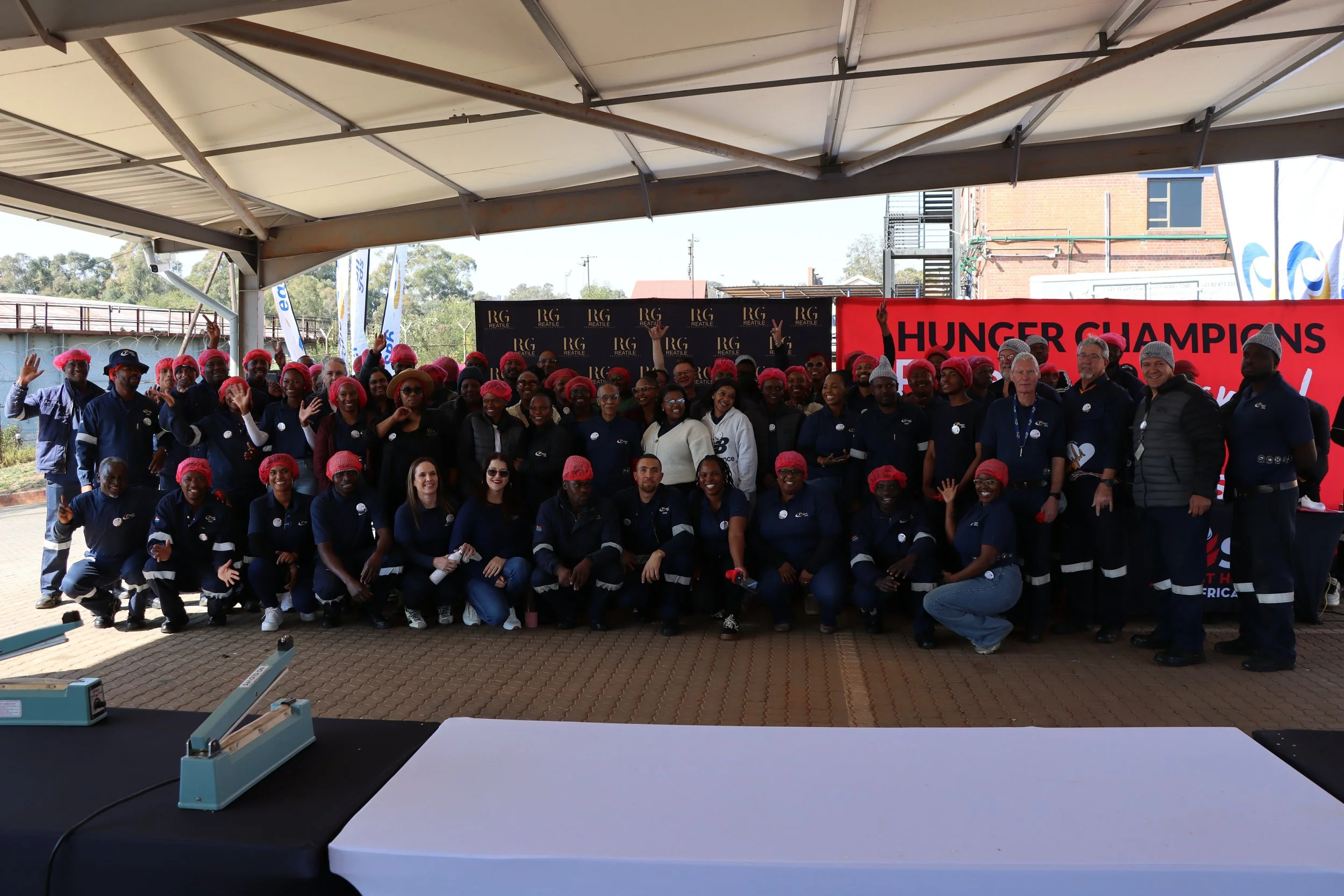Group of people wearing red hairnets standing under a tent, posing for a photo at an outdoor event with a banner that reads 'HUNGER CHAMPIONS' in the background.