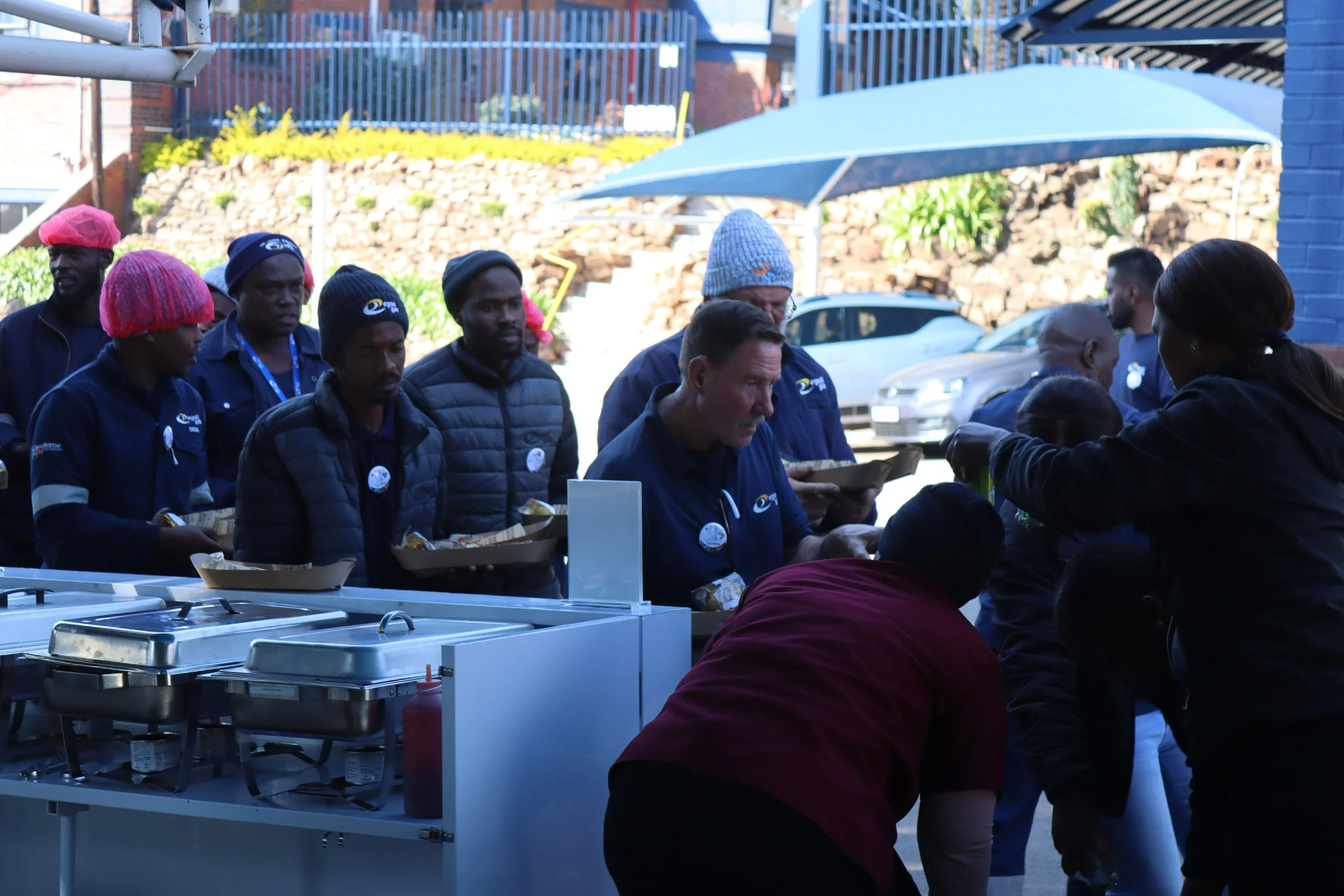 People in outdoor setting gathered around a food service counter, serving food to customers, with some wearing pink hairnets and navy uniforms, under blue umbrellas with a brick wall and parked cars in the background.