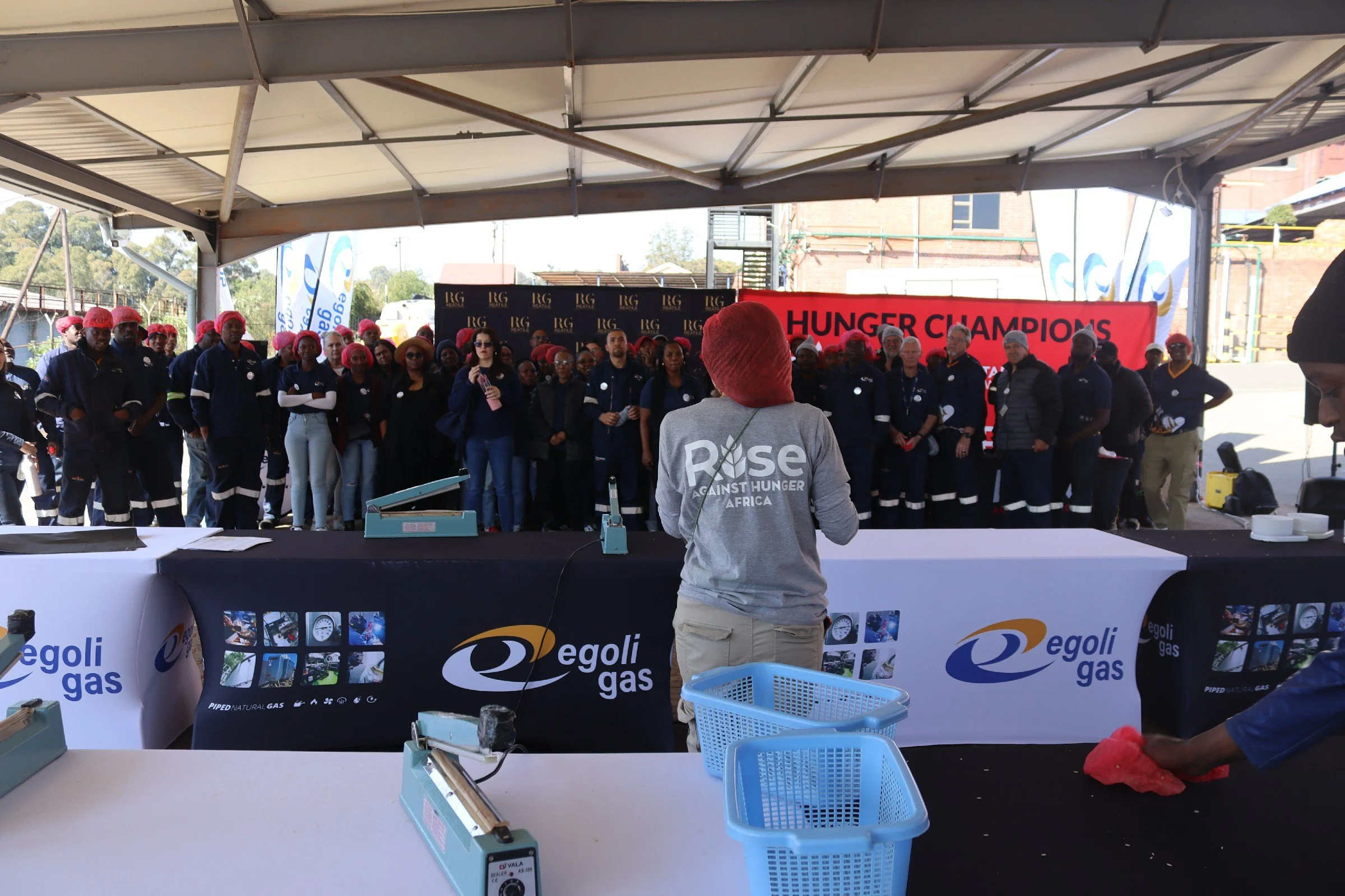 Group of people wearing red hairnets and dark uniforms gathered under a tent at an outdoor event, with banners and signs including 'Hunger Champions' and 'Egli Gas'. A woman with sunglasses and a dark jacket speaks into a microphone, facing the group