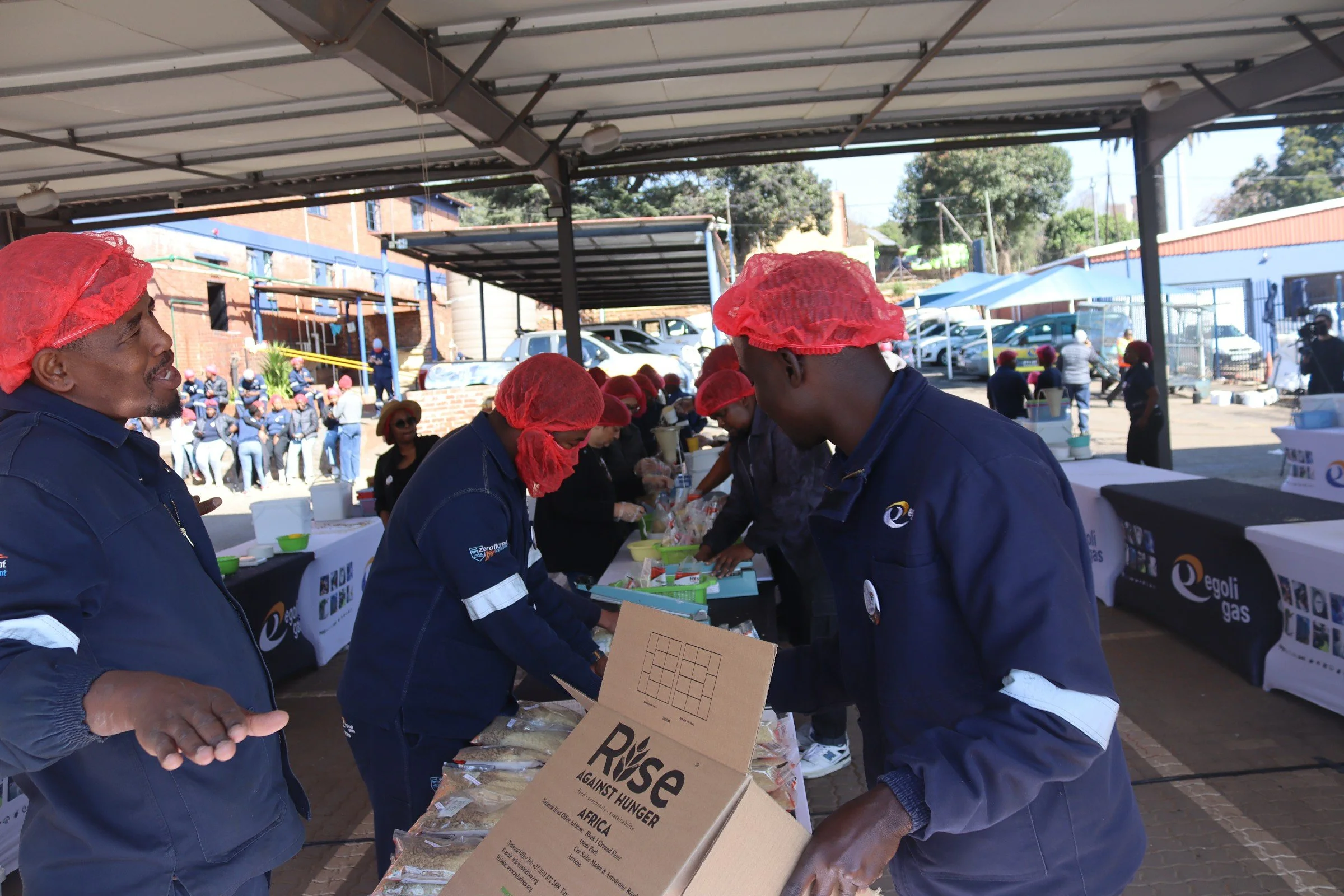 Group of people wearing red hairnets and navy blue uniforms standing at a table with food items, participating in a charity event outdoors. Some people are organizing and handing out food packages labeled with a hunger relief message, with a crowd an