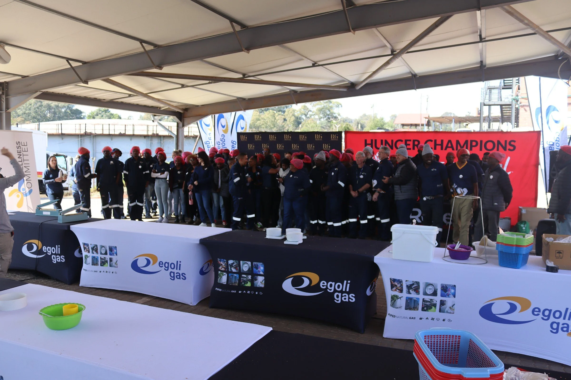 Group of people wearing dark uniforms and red hats standing under a tent at an event. There are banners with the logo 'egoli gas' and a red banner with text 'HUNGER CHAMPIONS'. Tables with bowls and baskets are in the foreground.
