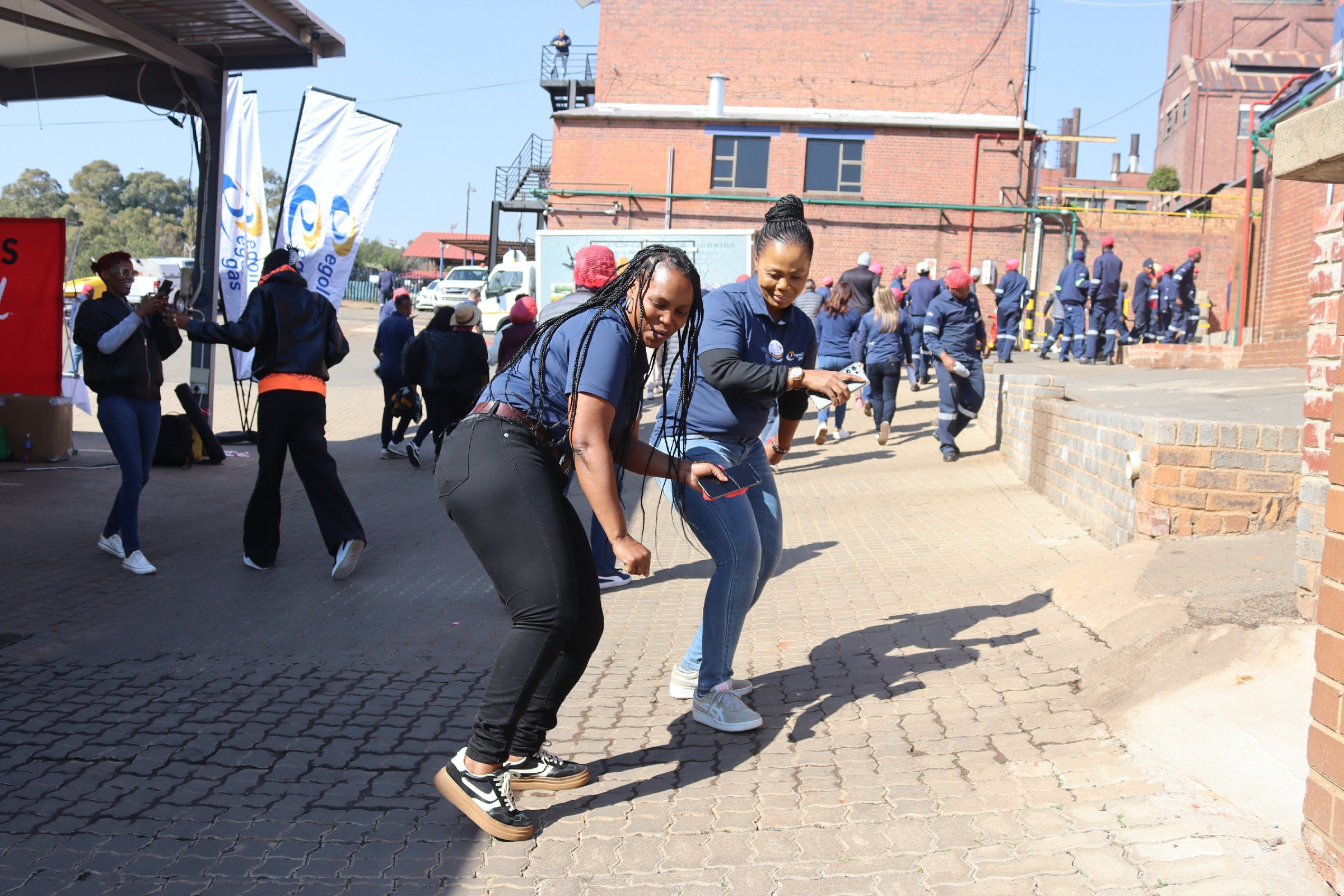 People dancing outdoors on a sunny day. Two women in the foreground are dancing, while others are walking or standing behind them. There are flags with logos, brick buildings, and a mural in the background.