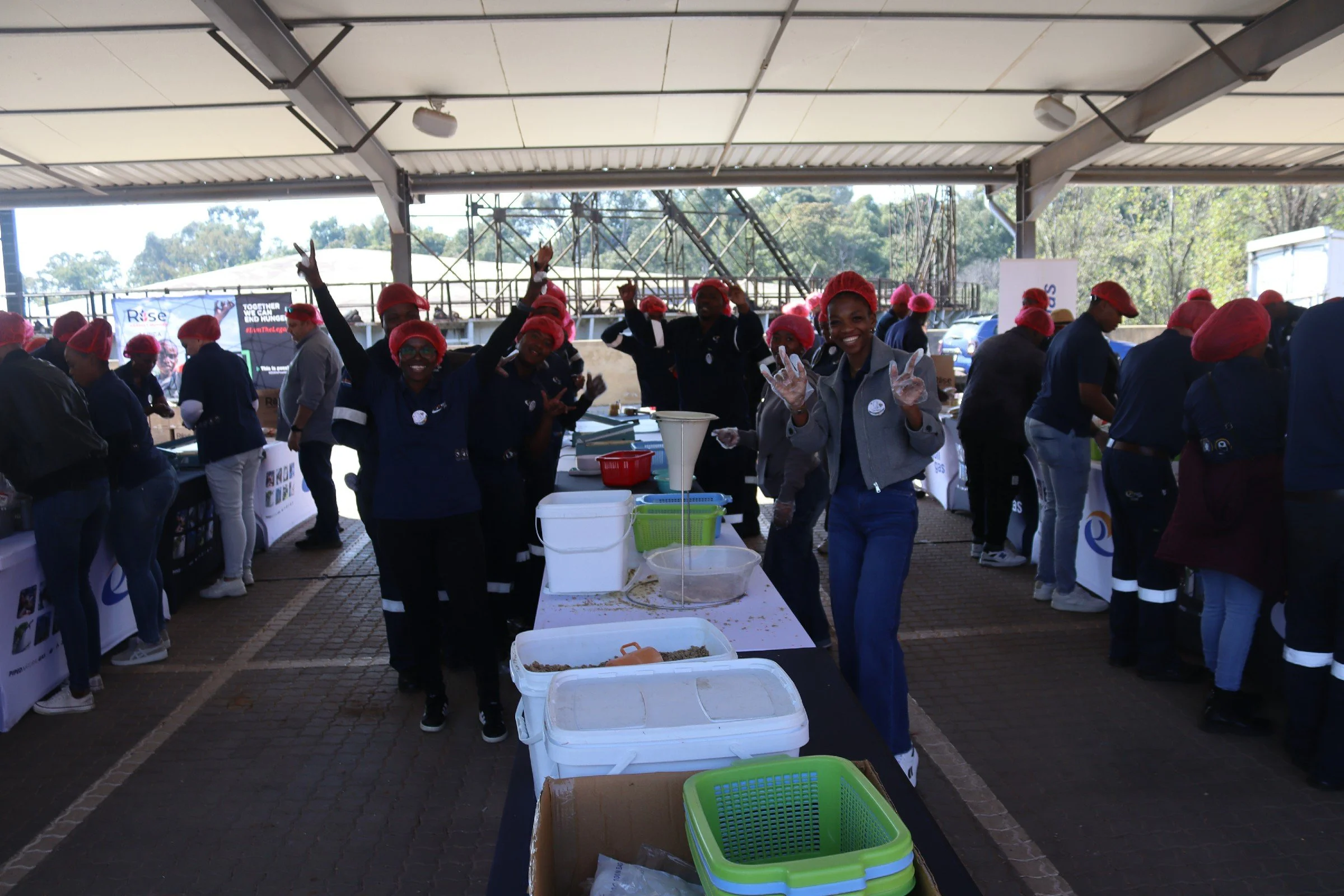 Group of people wearing red hair caps and gloves working together at a community event under a large shelter, with tables and containers filled with food or supplies.