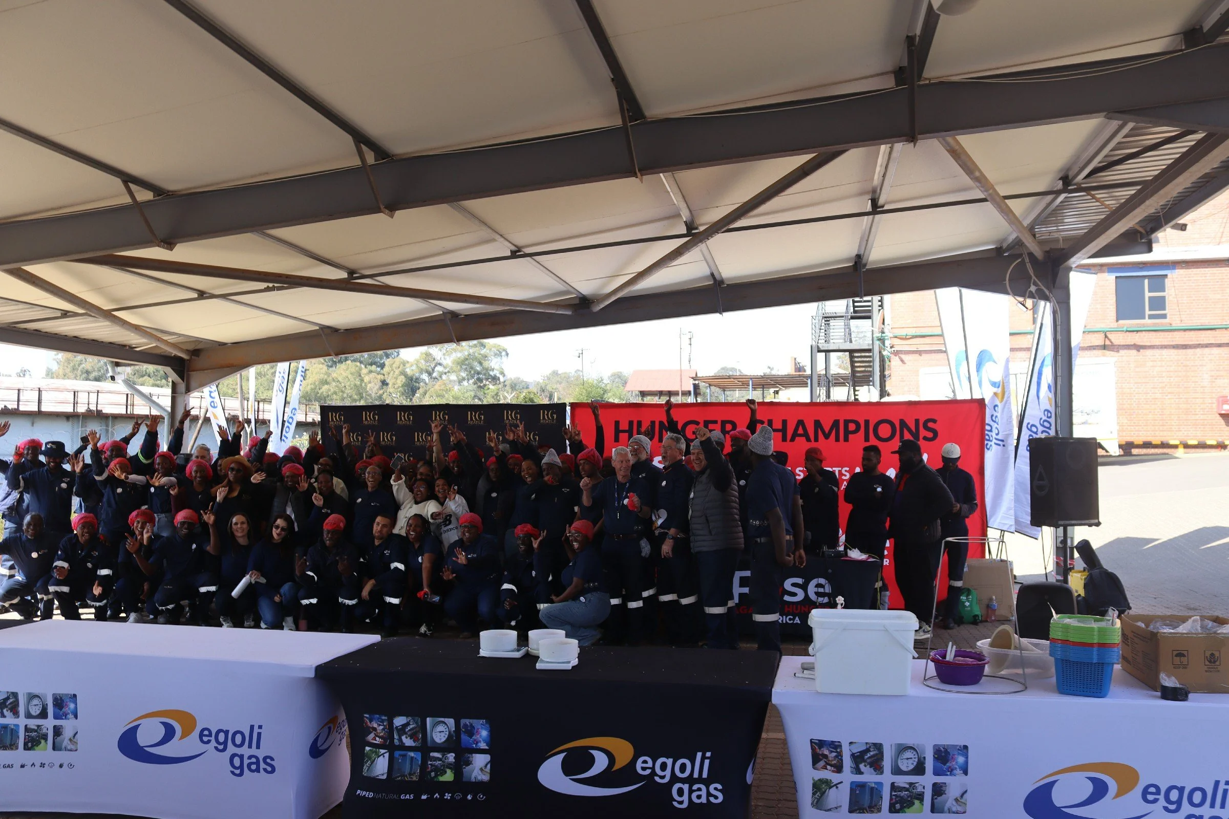 Group of people gathered under a tent, holding up their hands in a celebratory manner, with banners and tables in the foreground featuring Egoli Gas branding, during an outdoor event.