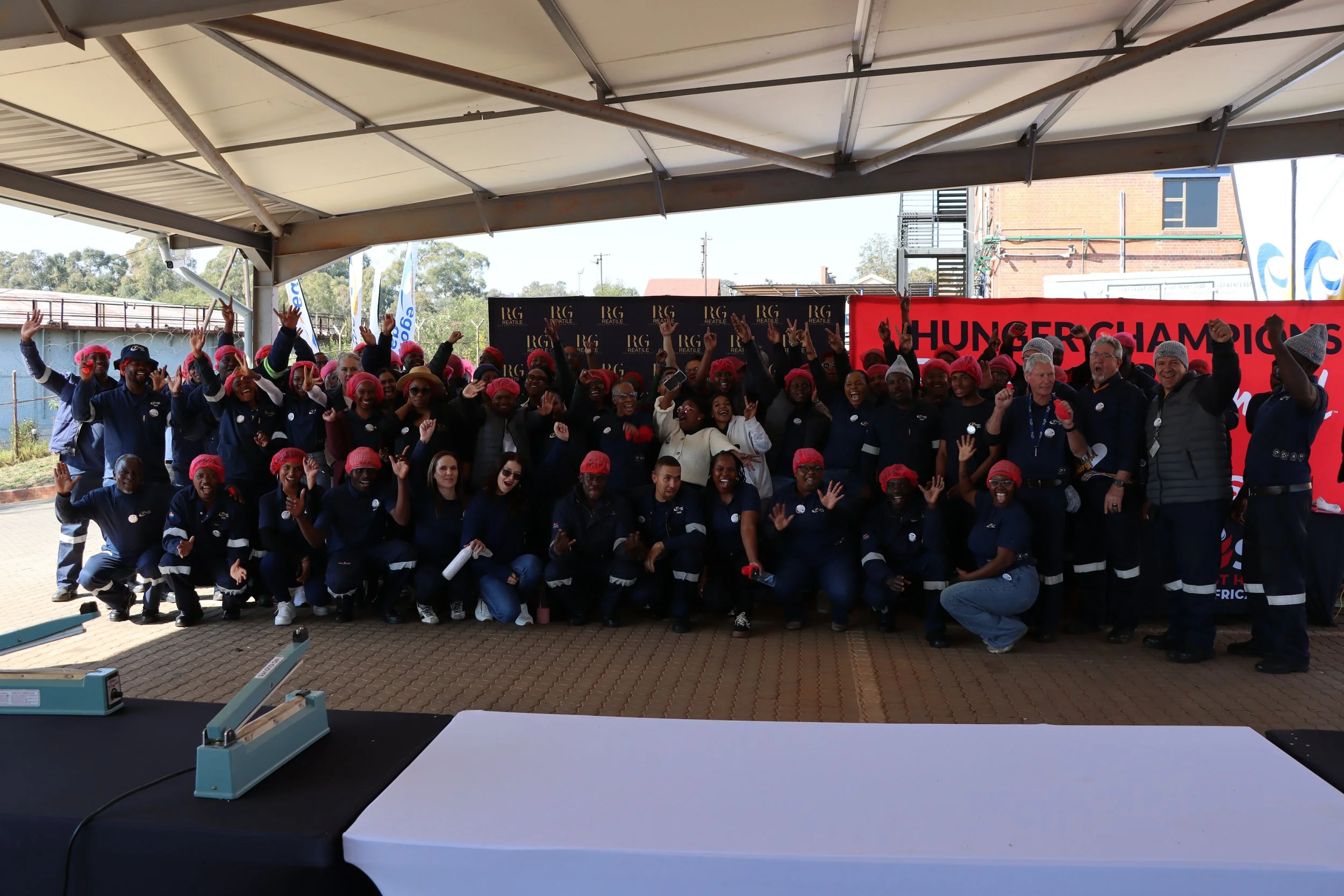 A large group of people, many of whom are wearing pink hairnets, poses under a covered outdoor area, celebrating. They are smiling and waving, with a backdrop that includes a red banner with partial text '#CHAMPIONS' and black panels with gold letter