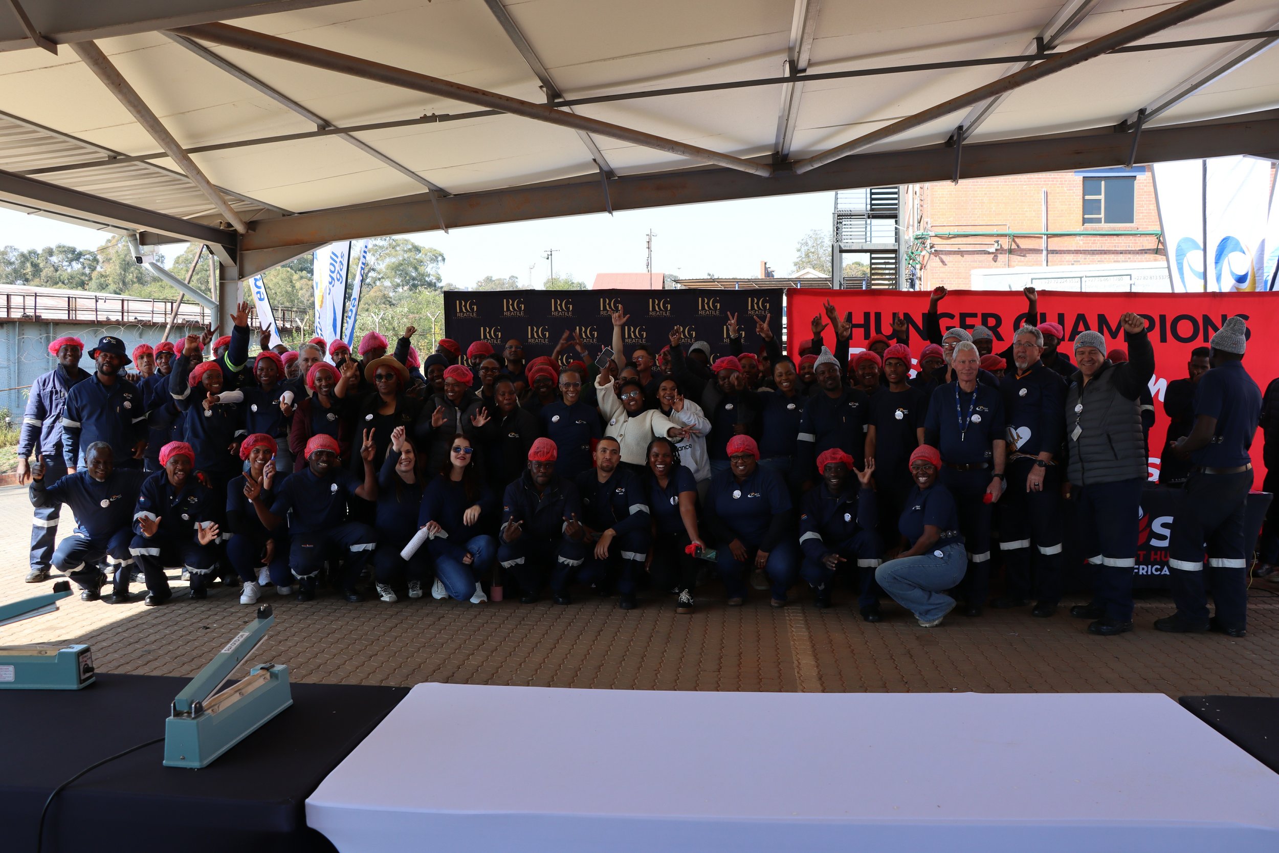 Large group of people wearing red hairnets and dark uniforms, standing under a tent outdoors, celebrating with raised fists, with a banner in the background that reads 'HUNGER CHAMPIONS'.