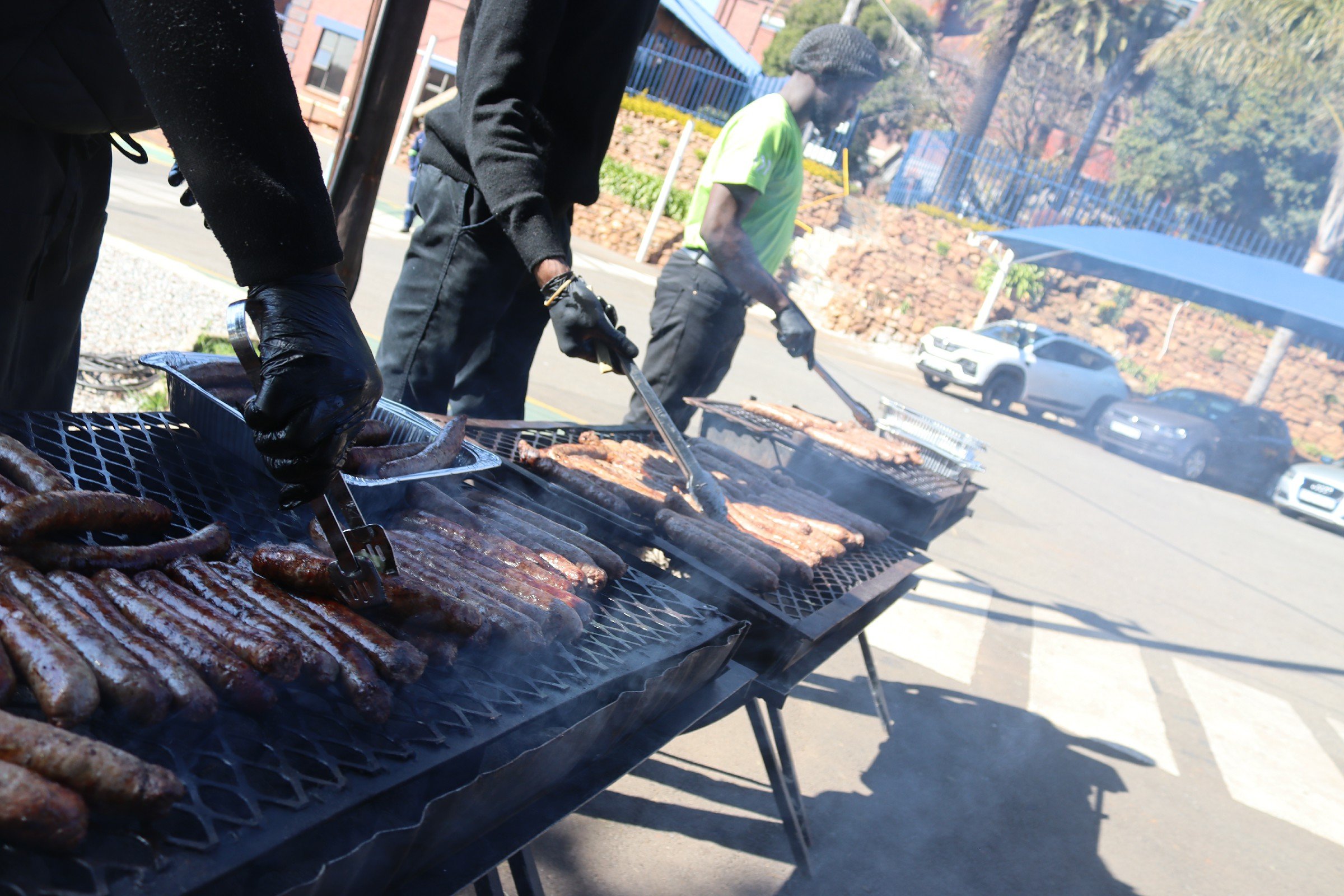 People cooking sausages and ribs on a grill outdoors, with parking and trees in the background.
