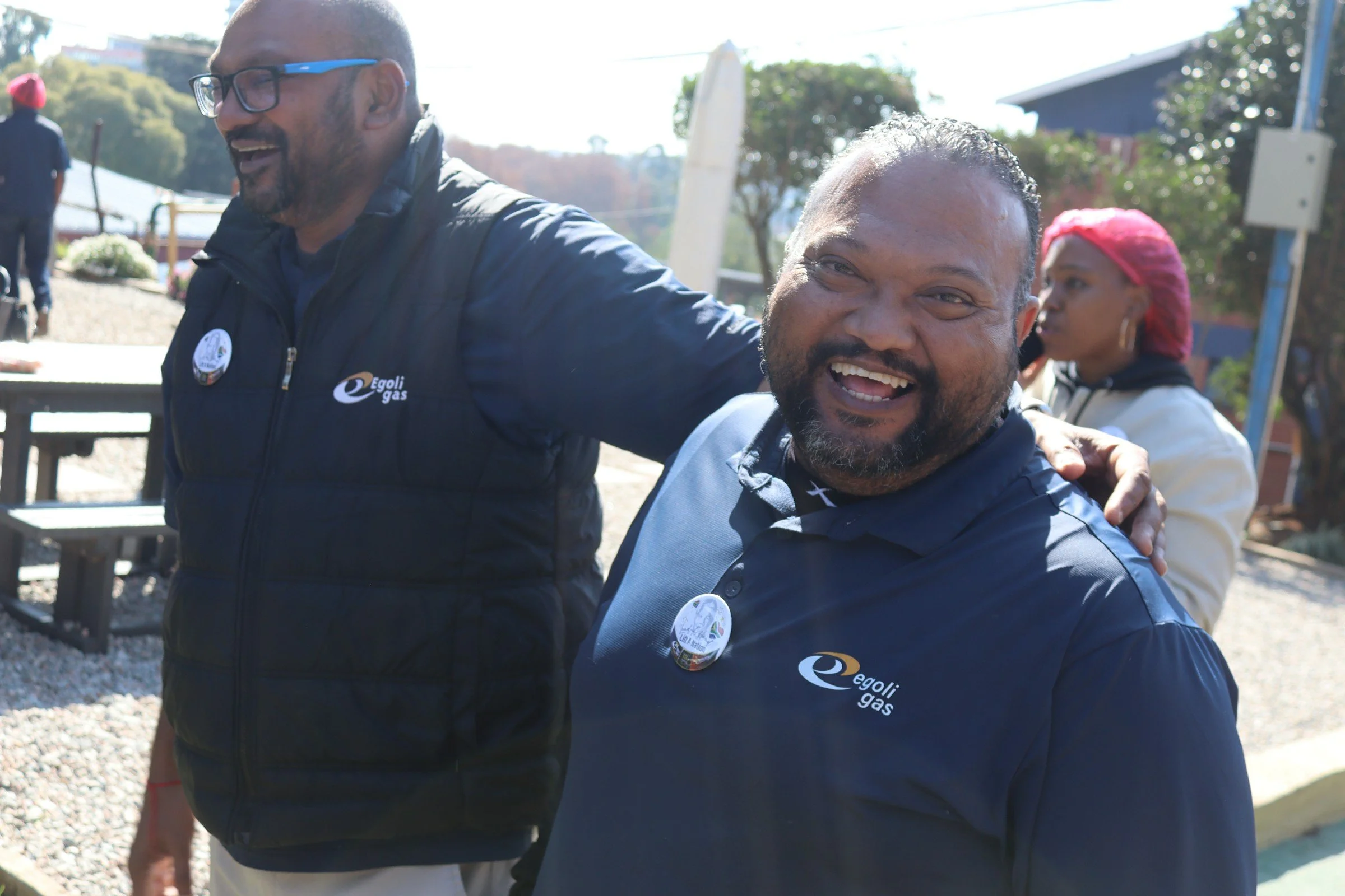 Two men smiling and embracing outdoors, with trees and a building in the background. Both wearing jackets with the 'Egoli Gas' logo and buttons, indicating a possible event or celebration.