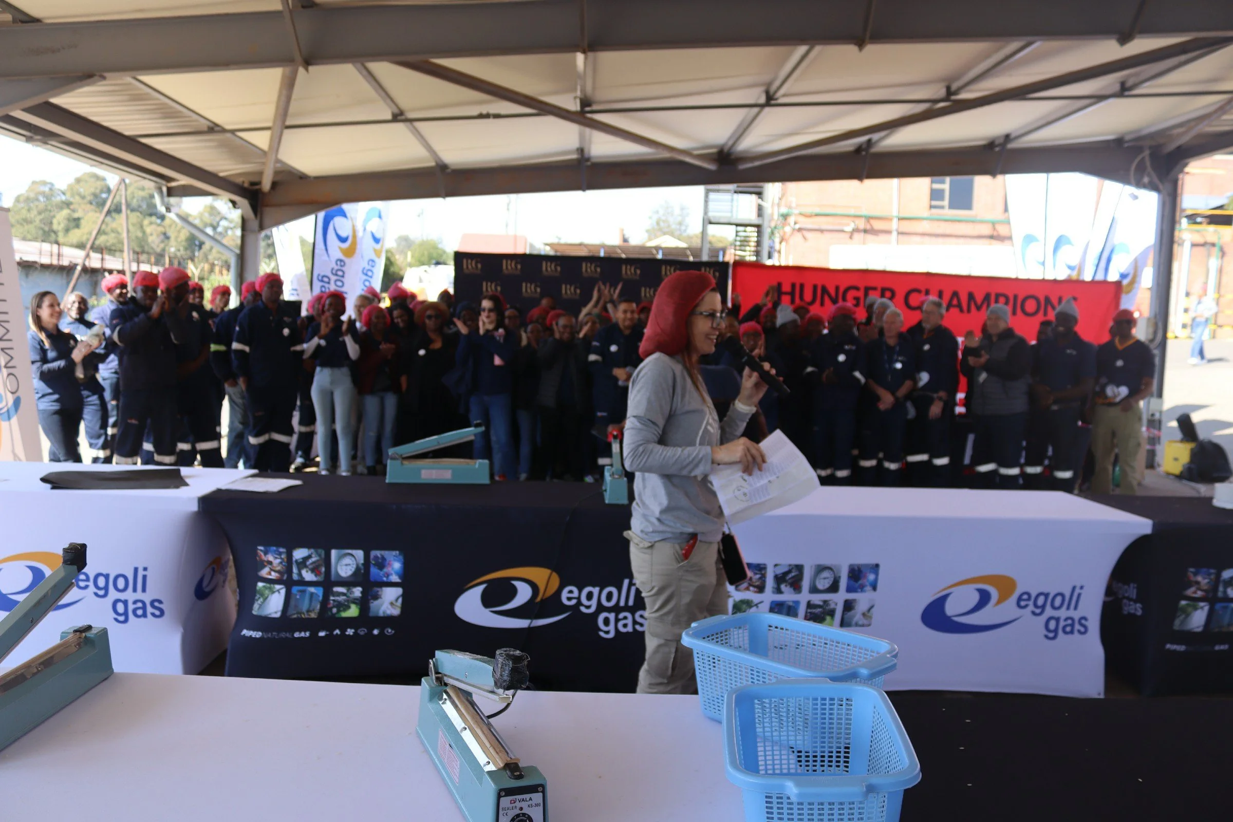 A woman in glasses and a gray jacket holds a microphone and a paper in front of a group of people wearing shirts and red head coverings, standing under an outdoor shelter with banners and equipment.