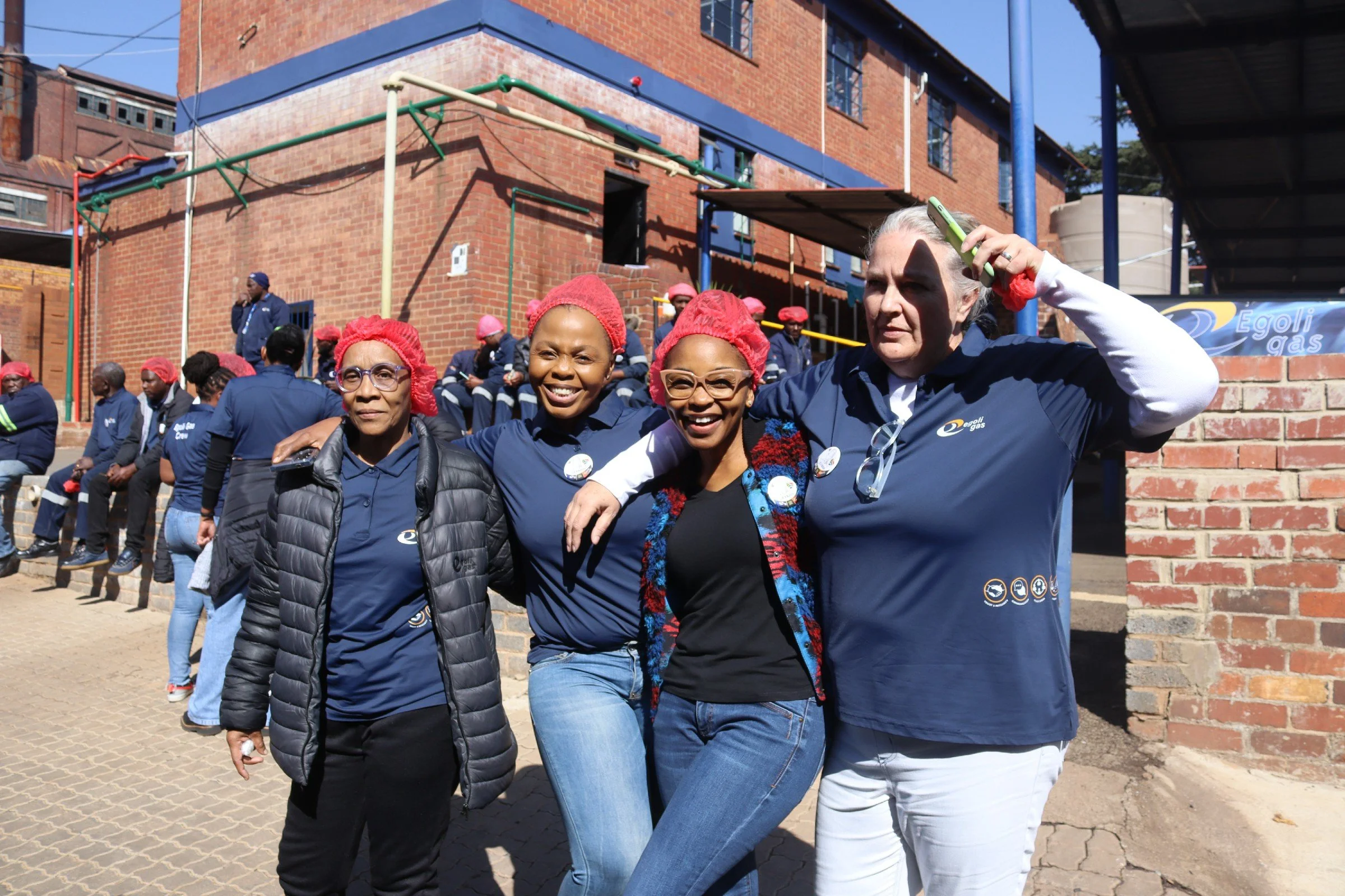 A group of smiling women and a man wearing navy blue shirts, glasses, and red hairnets, standing together outdoors in front of a brick building, with others sitting and standing in the background, some also with red hairnets.