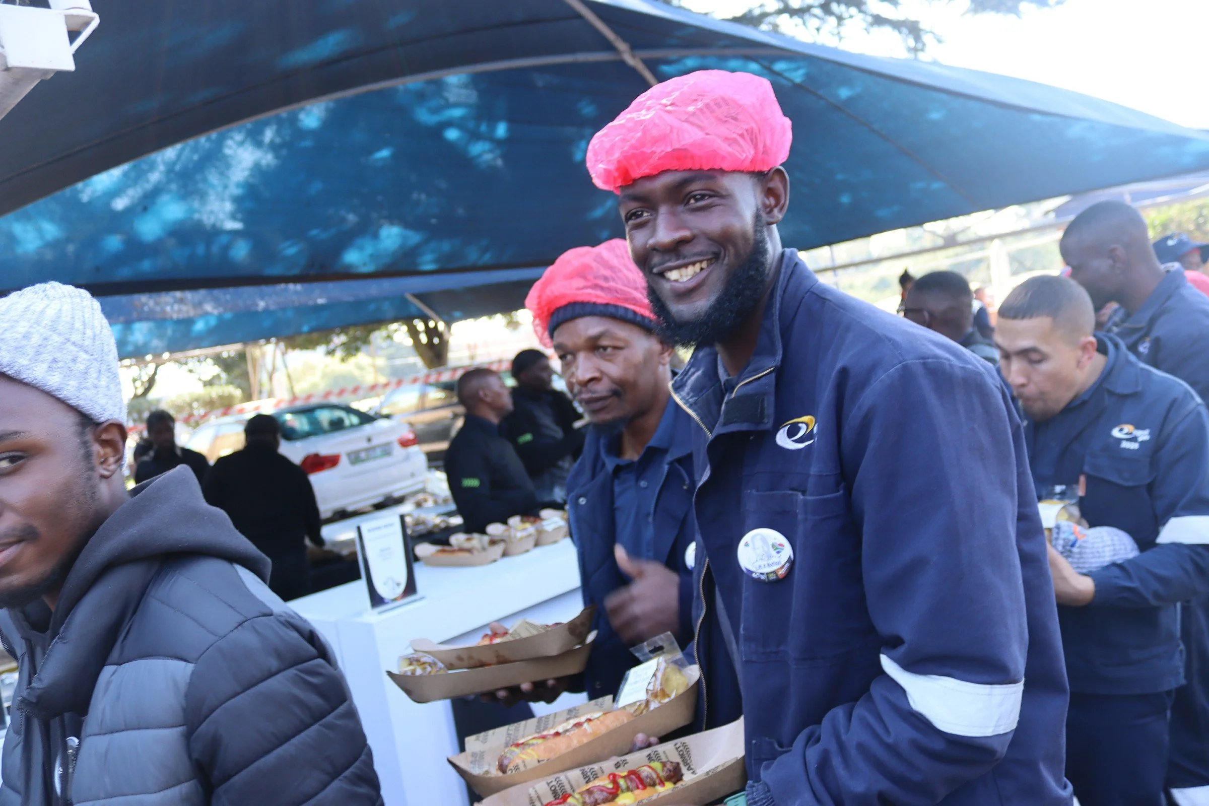 A group of people gathered outdoors under a canopy, with a man in the center smiling, wearing a pink hairnet, and selling snacks from paper bags.