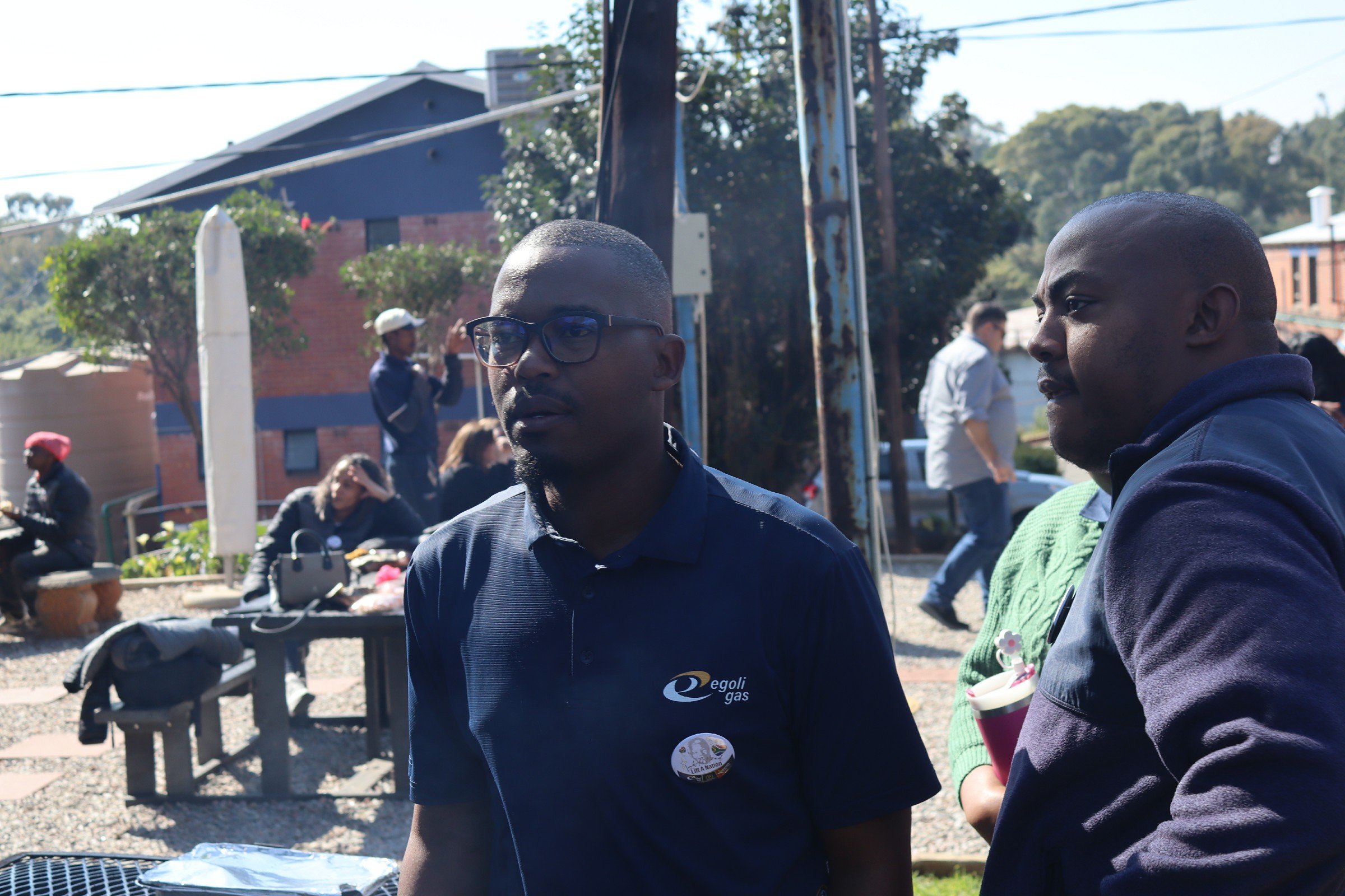 Two men with dark skin and short hair standing outdoors, engaged in conversation. One wears glasses and a dark shirt with a logo, the other wears a green sweater over a shirt. In the background, there are other people, trees, and buildings, with a su