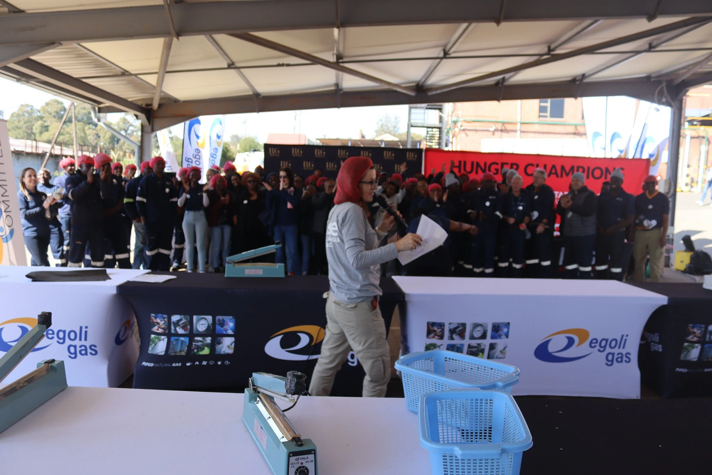A group of people wearing red hair coverings and dark uniforms gathered under a tent for a ceremony or event. A woman is speaking into a microphone in front of the group, with banners and signs in the background.