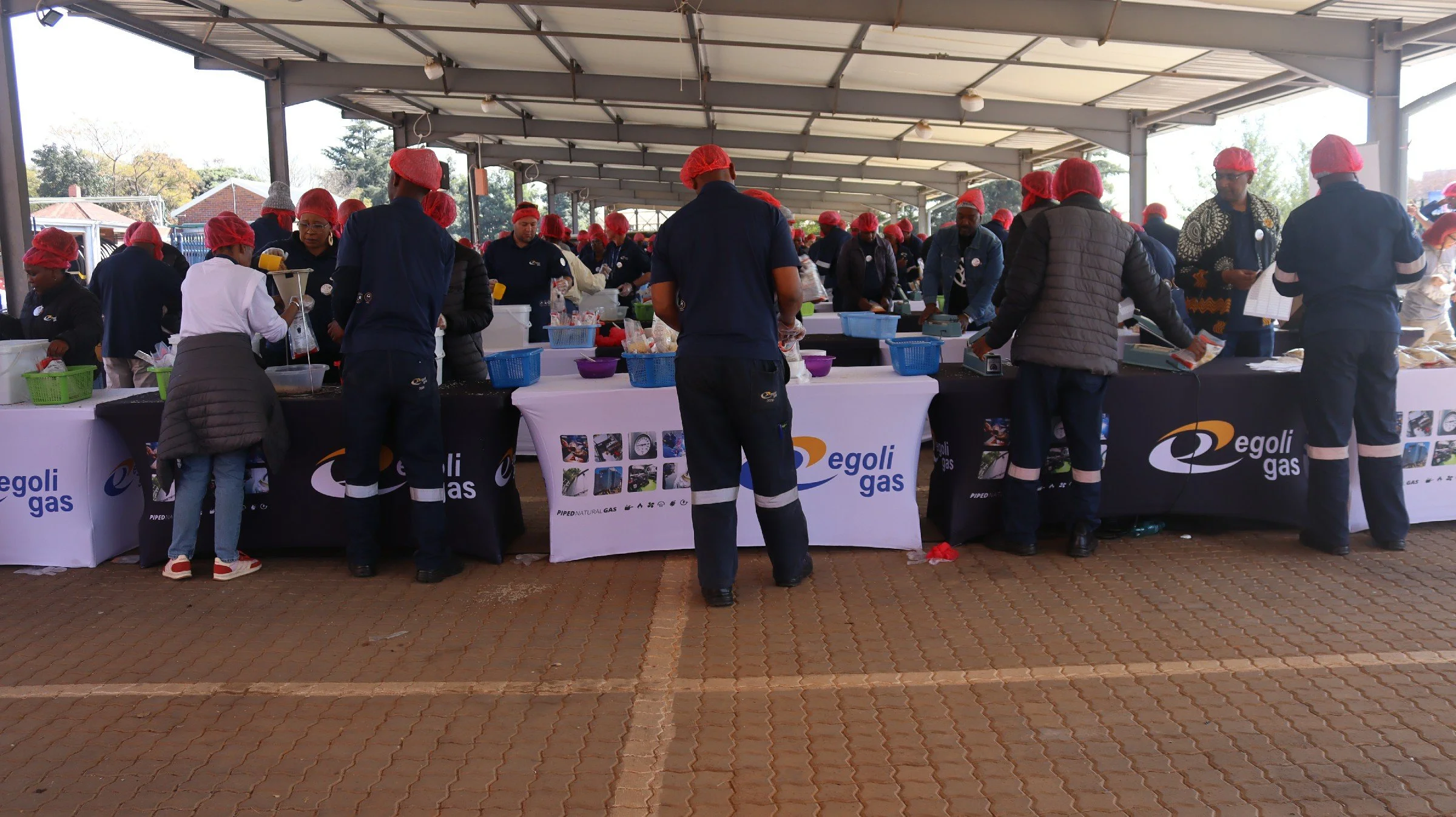 People working at an outdoor market stall under a roof, wearing red hairnets and dark clothing, with promotional banners for Egoli Gas. The stall has various products and a scale, with several customers and staff interacting.