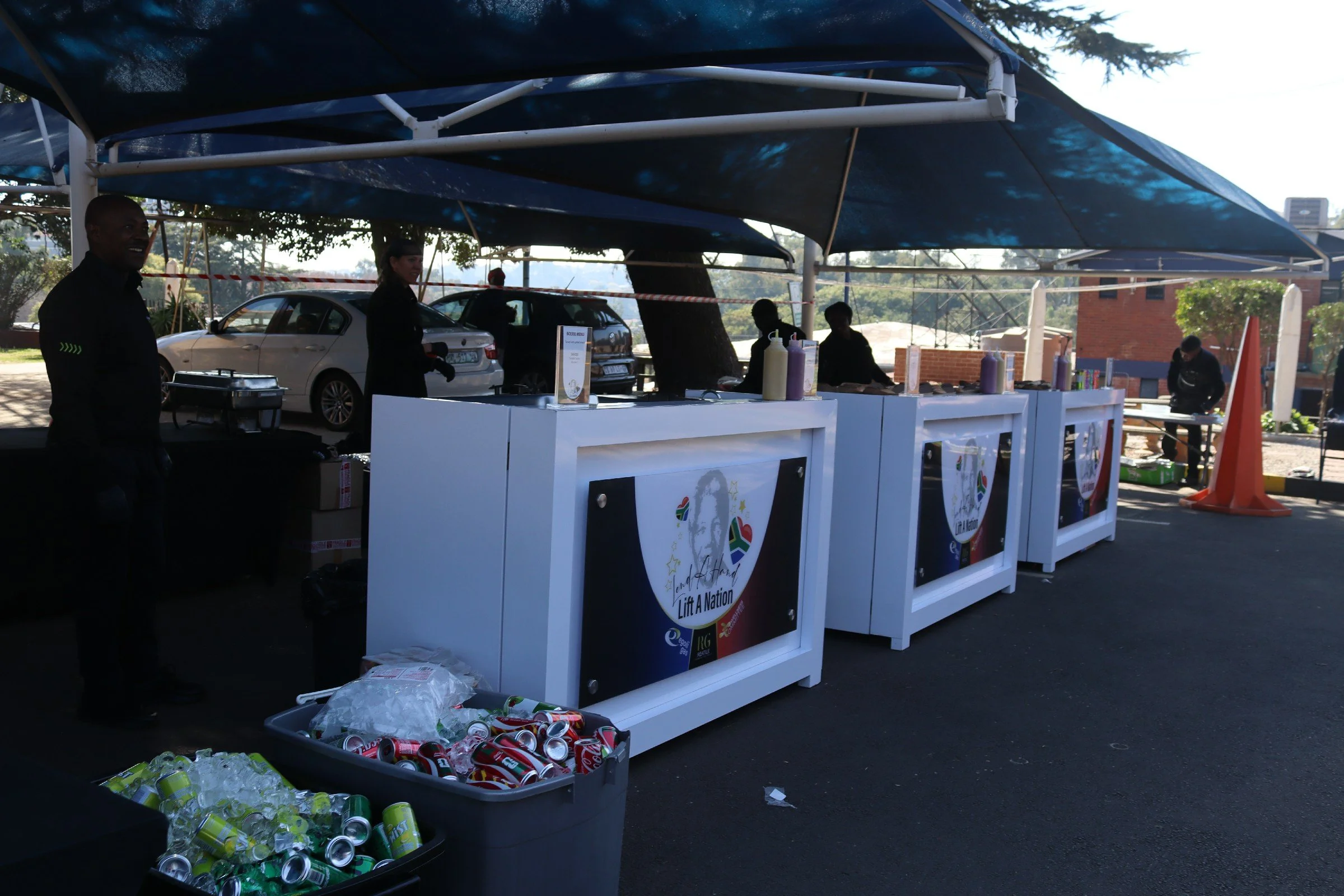 A donation or recycling booth with signs that say 'Lift A Nation', under large blue shade sails, with cans and bottles in bins and people managing the booth