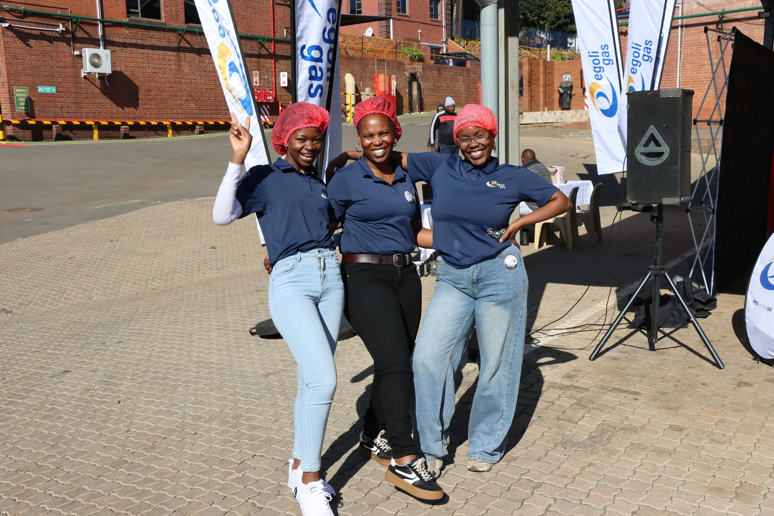 Three women wearing blue shirts and red hair caps smiling and posing together at an outdoor event with flags that say "egoli gas" and a brick wall background.