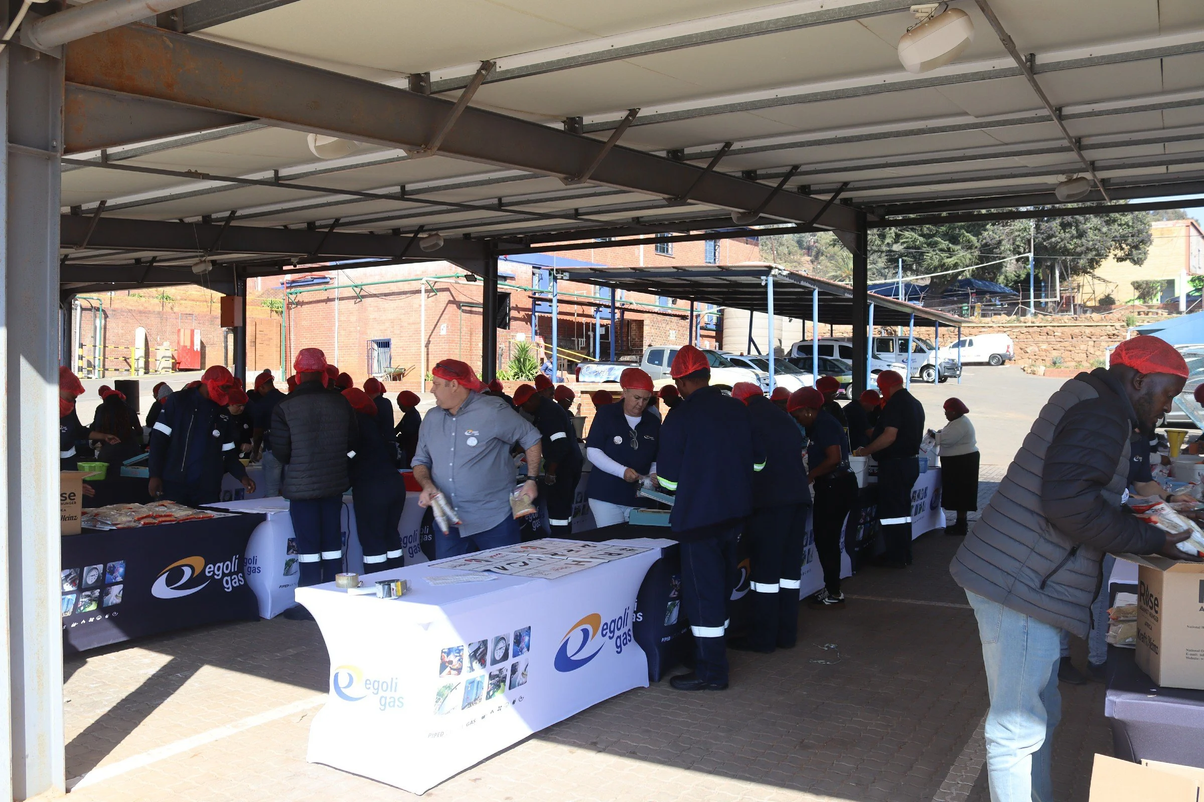 People wearing uniforms and red hats working at tables under a covered outdoor area, with cars parked in the background.