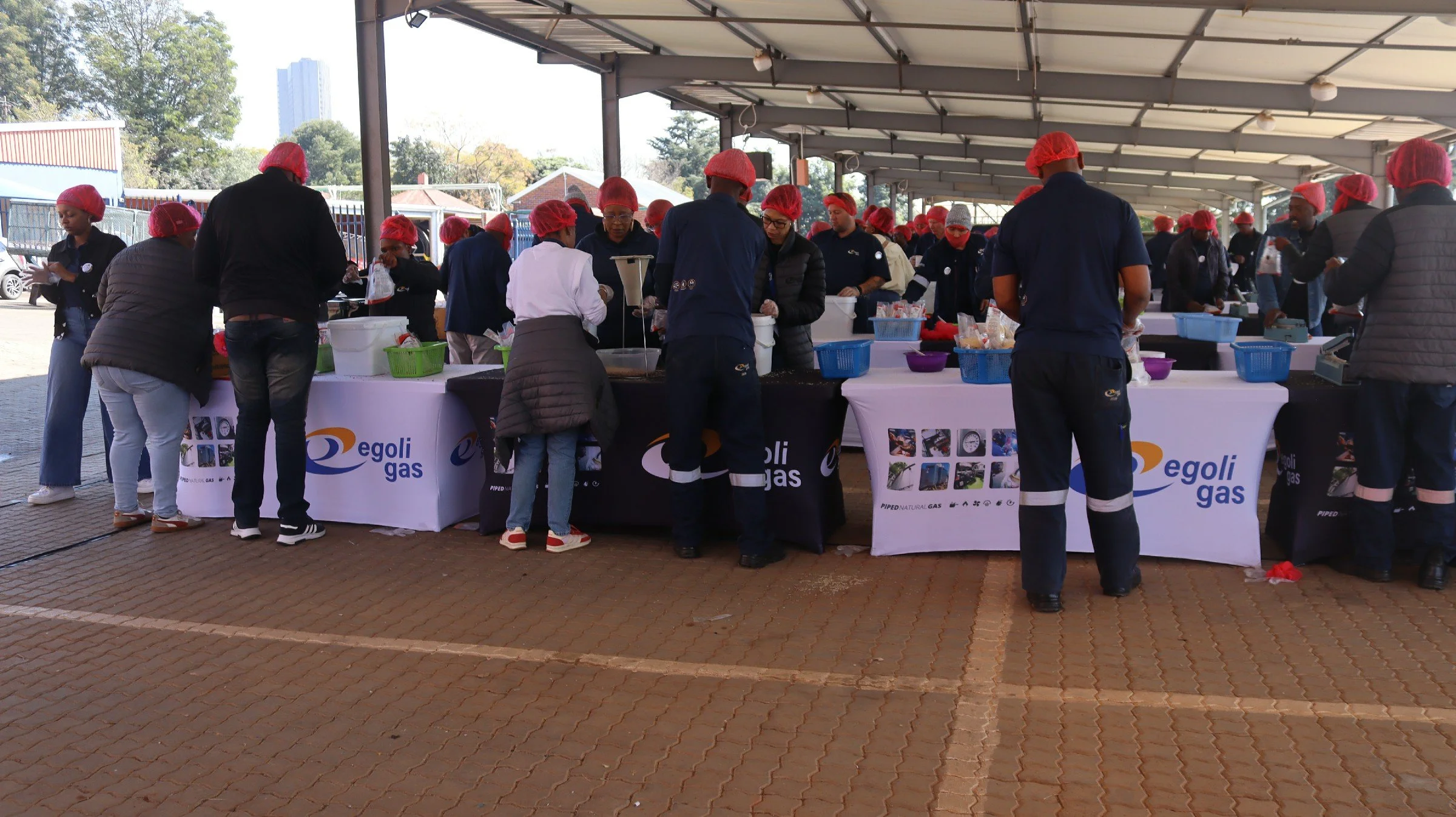 People wearing red hairnets and gloves standing behind tables at an outdoor event, serving food or distributing items, with banners displaying the 'Egoli Gas' logo.