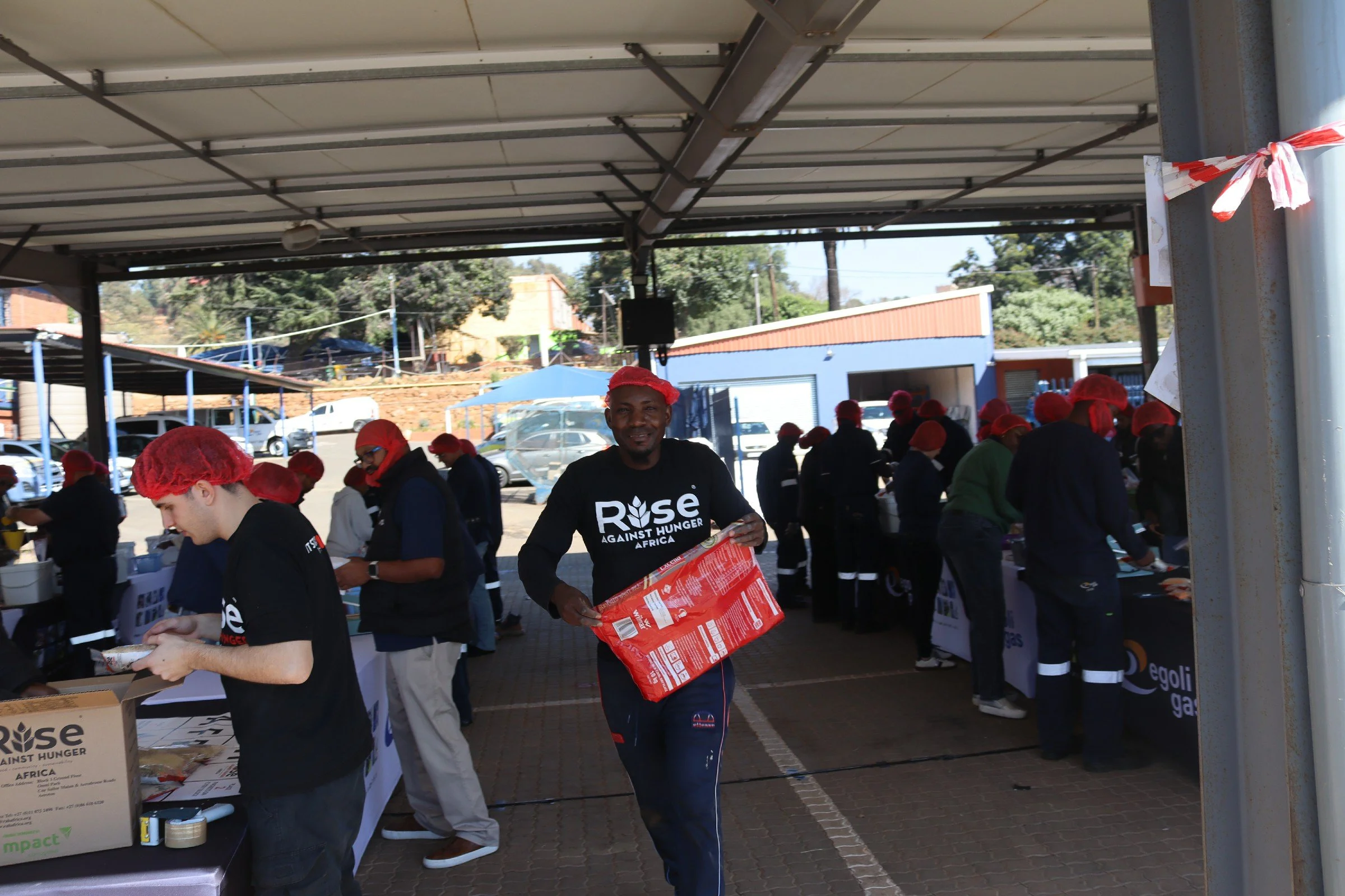 A group of volunteers under a parking lot shelter wearing red hairnets and black shirts with white text, distributing food or supplies for a charity event.