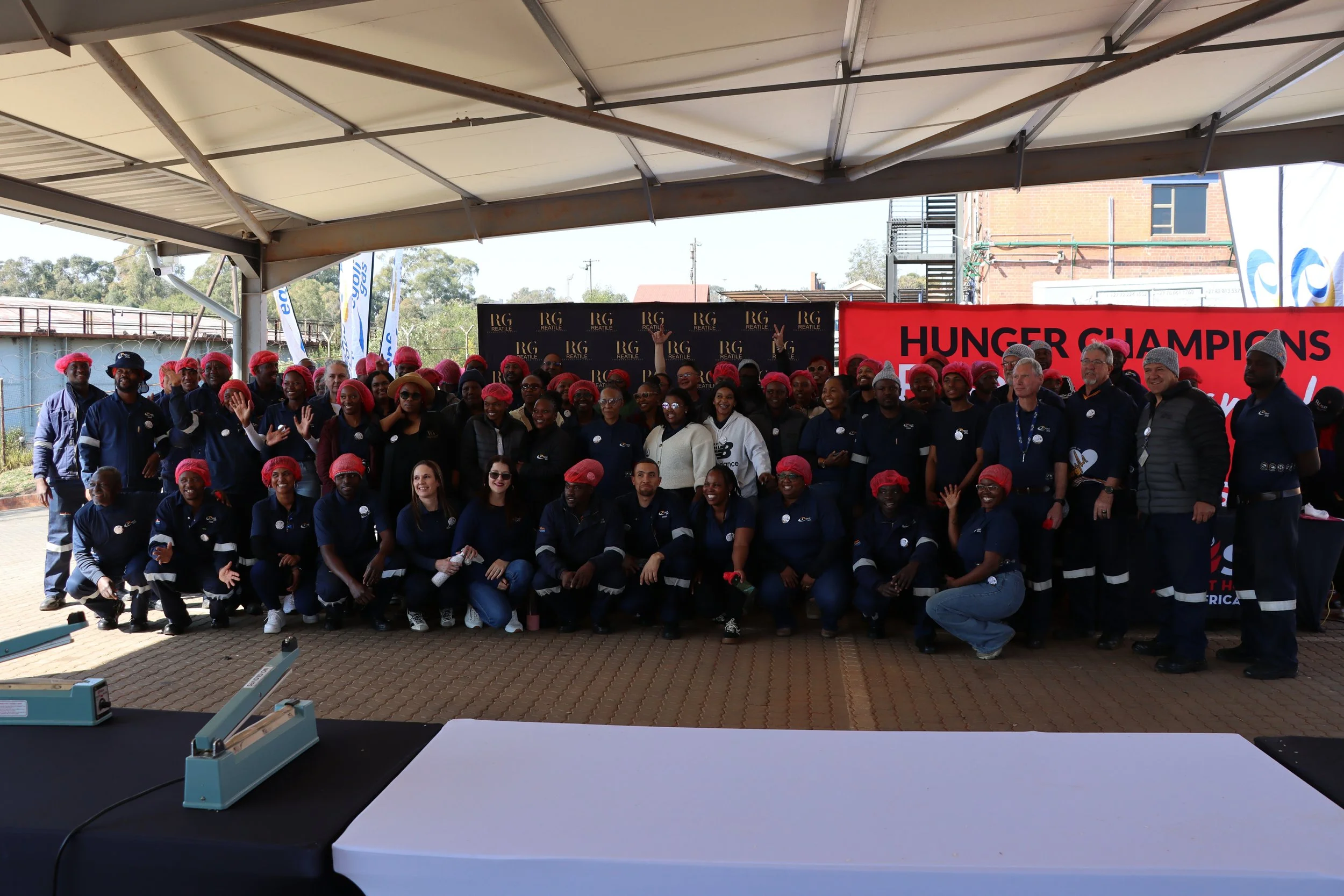 Group of people gathered under an outdoor shelter, wearing red hairnets and dark blue uniforms, smiling and posing for a photo with a red banner that reads "HUNGER CHAMPIONS."