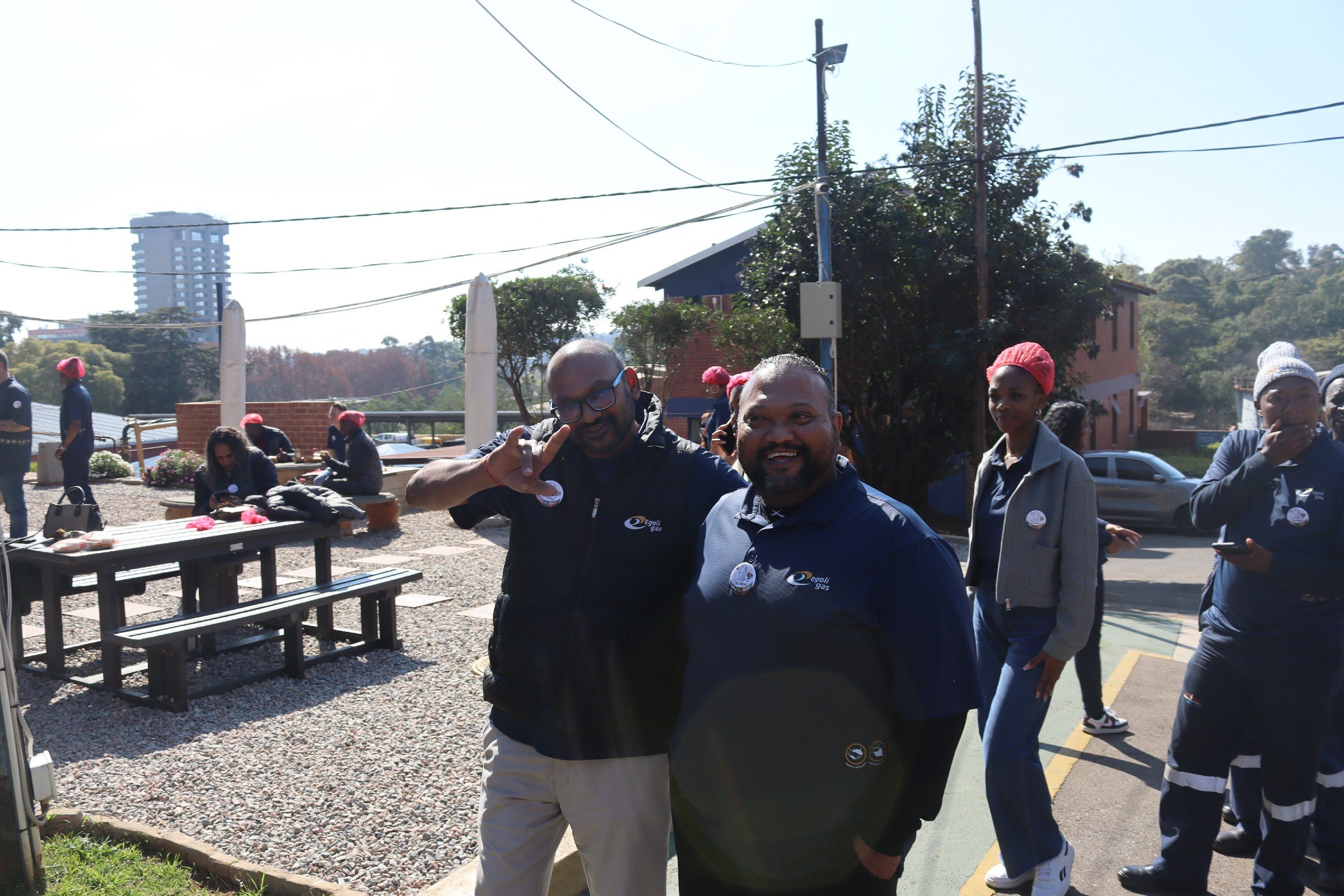 Two smiling men standing outdoors among a group of people, with some wearing red hats, near benches and trees, in what appears to be a sunny day at a community event or gathering.