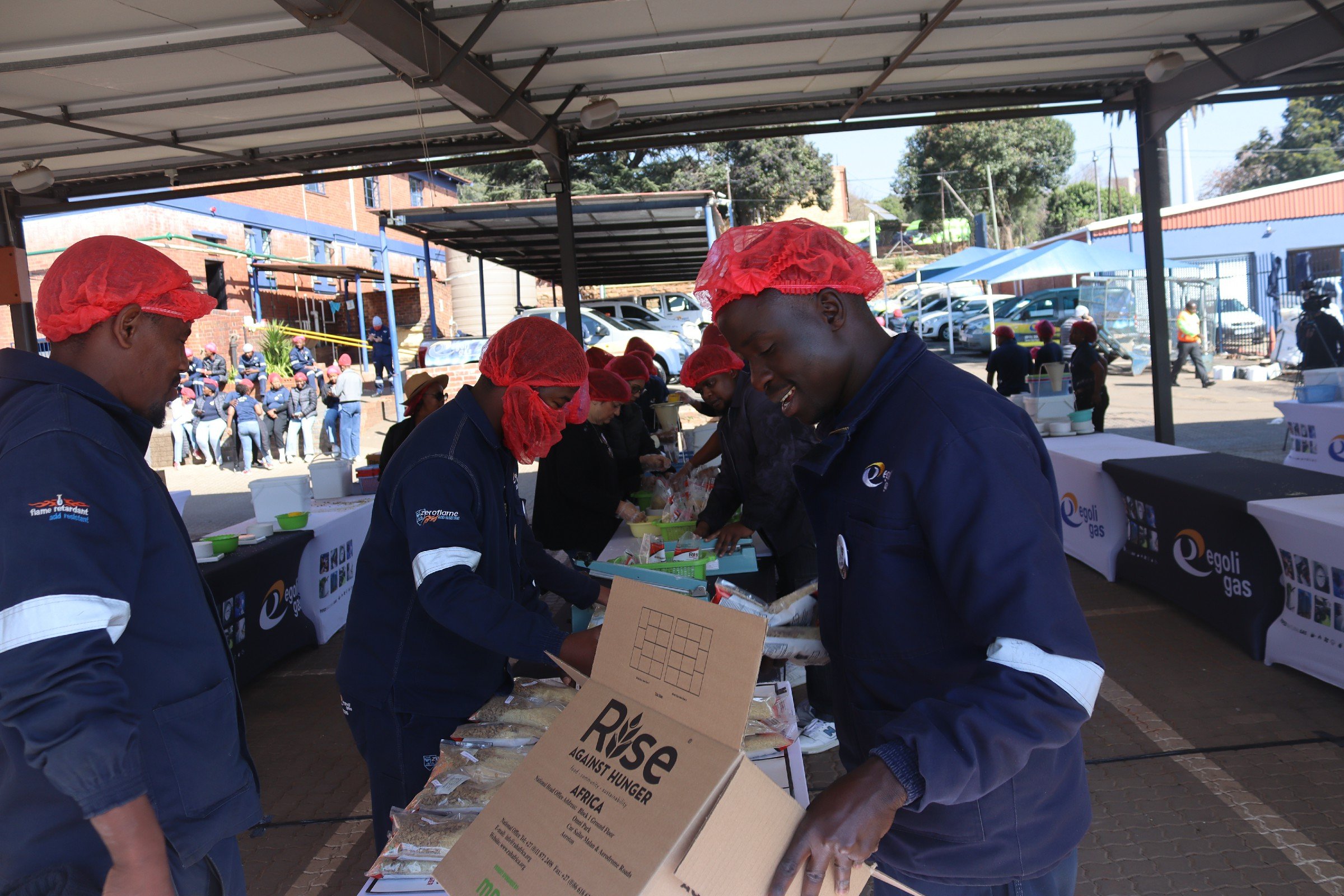 People wearing red hair covers, working at an outdoor food stall during a charity event, with a crowd and cars in the background.
