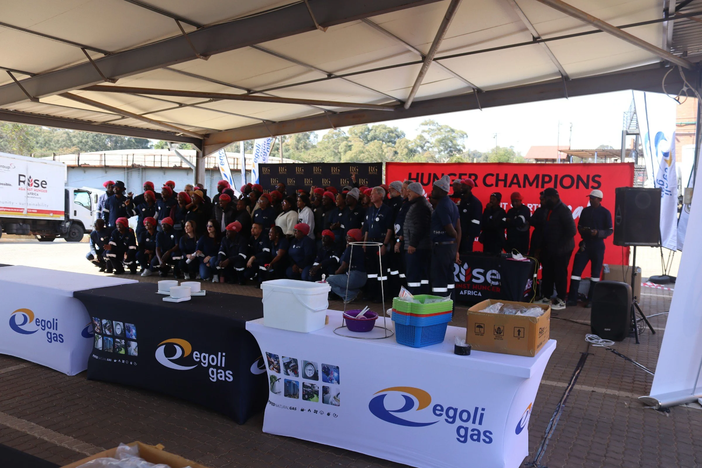 A group of people wearing matching uniforms and red hats standing on a stage at an outdoor event with banners and tables displaying logos, including Egoli Gas and Hunger Champions.