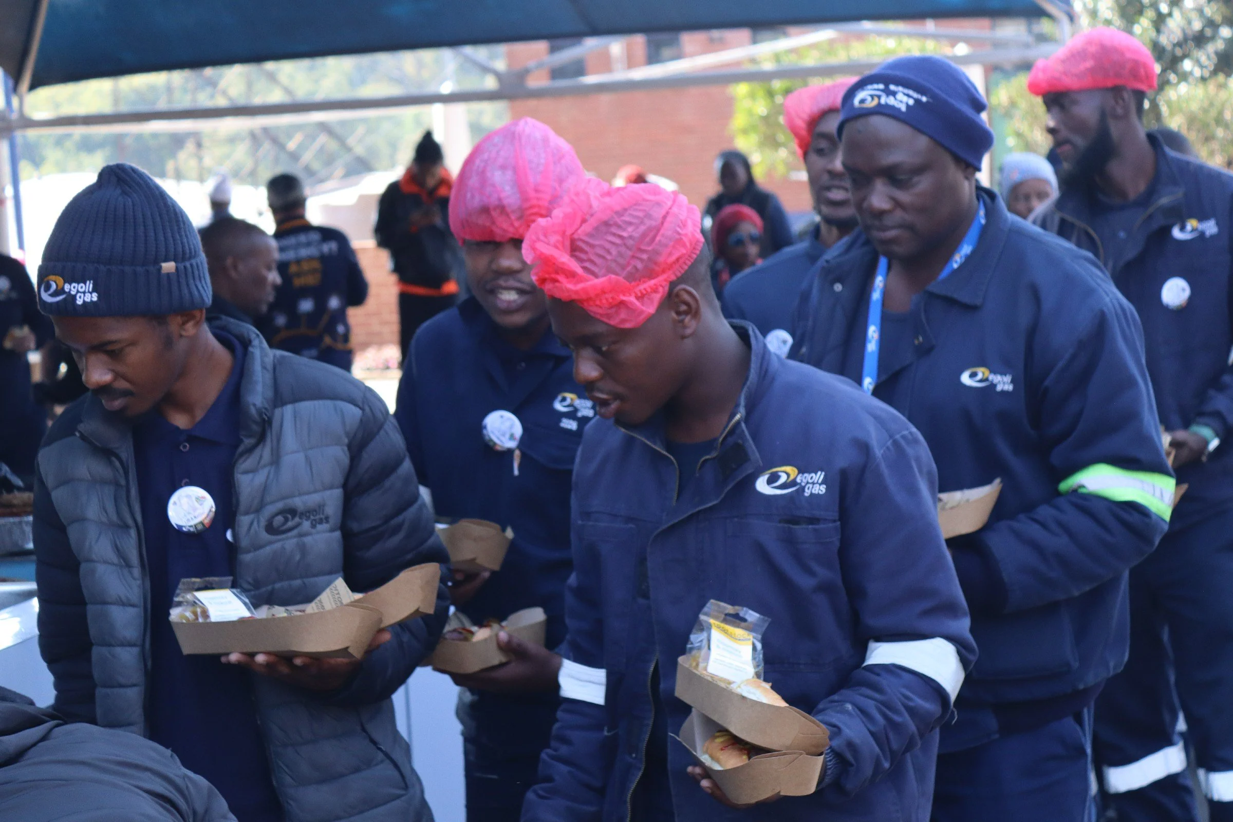 Group of people wearing blue jackets and pink hairnets standing outdoors, holding food trays during a community event.