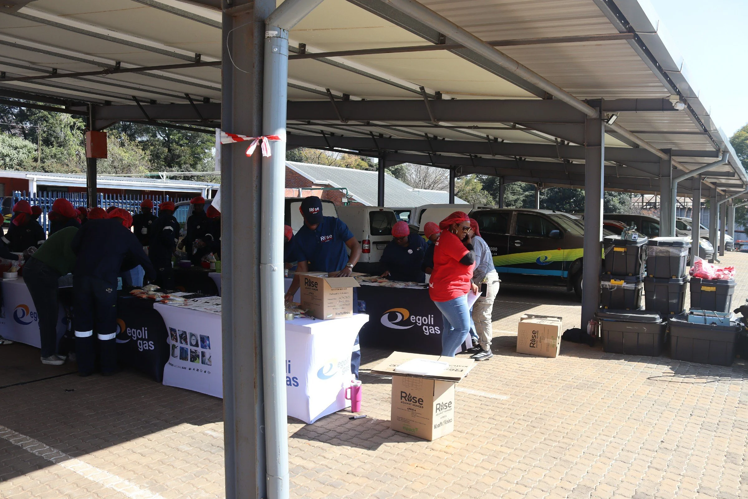 People wearing red hats working at tables under a metal carport during daytime, with boxes and banners displaying the 'Egoli Gas' logo.