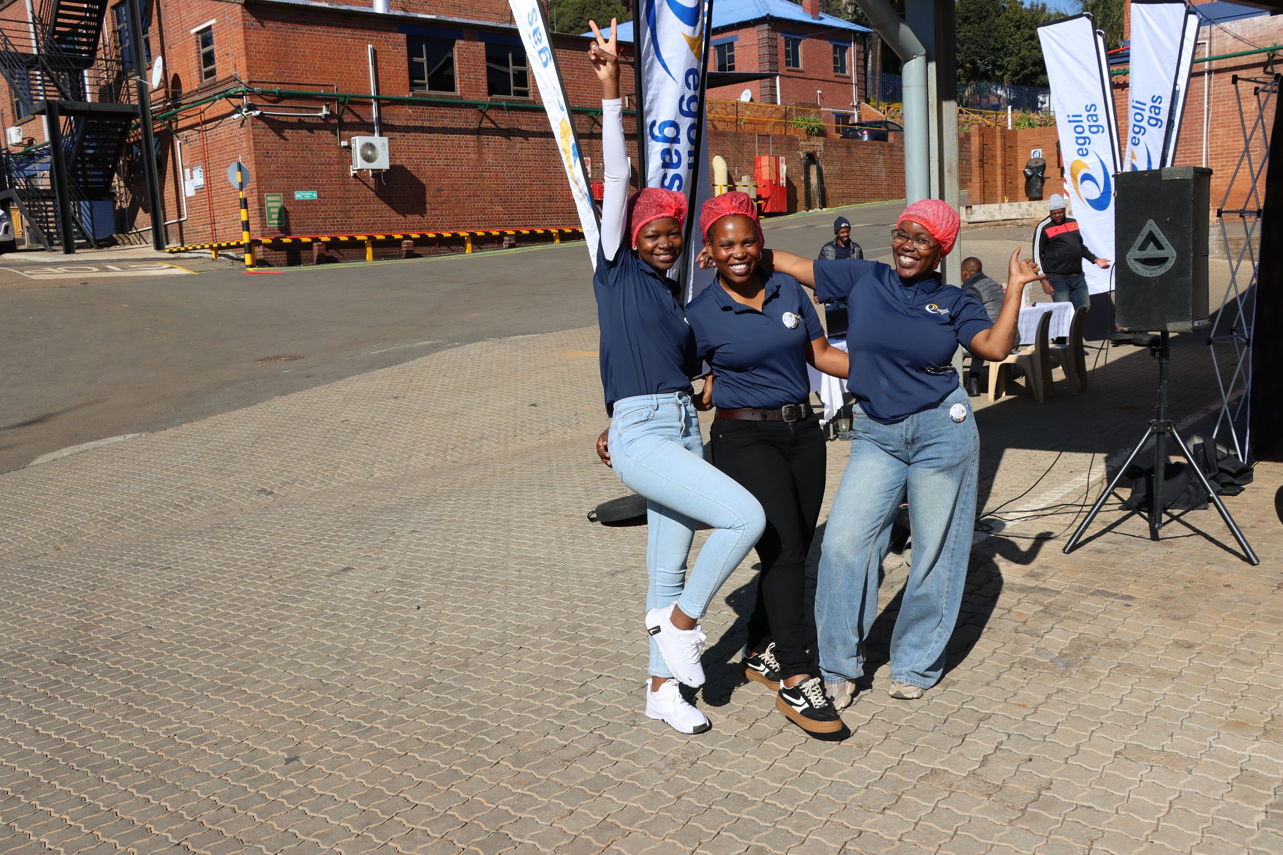 Three women wearing navy polo shirts, jeans, and red hair covers, are smiling and posing happily outdoors, with two raising their hands making peace signs. They are standing on a paved surface near banners and event equipment, with a brick building i