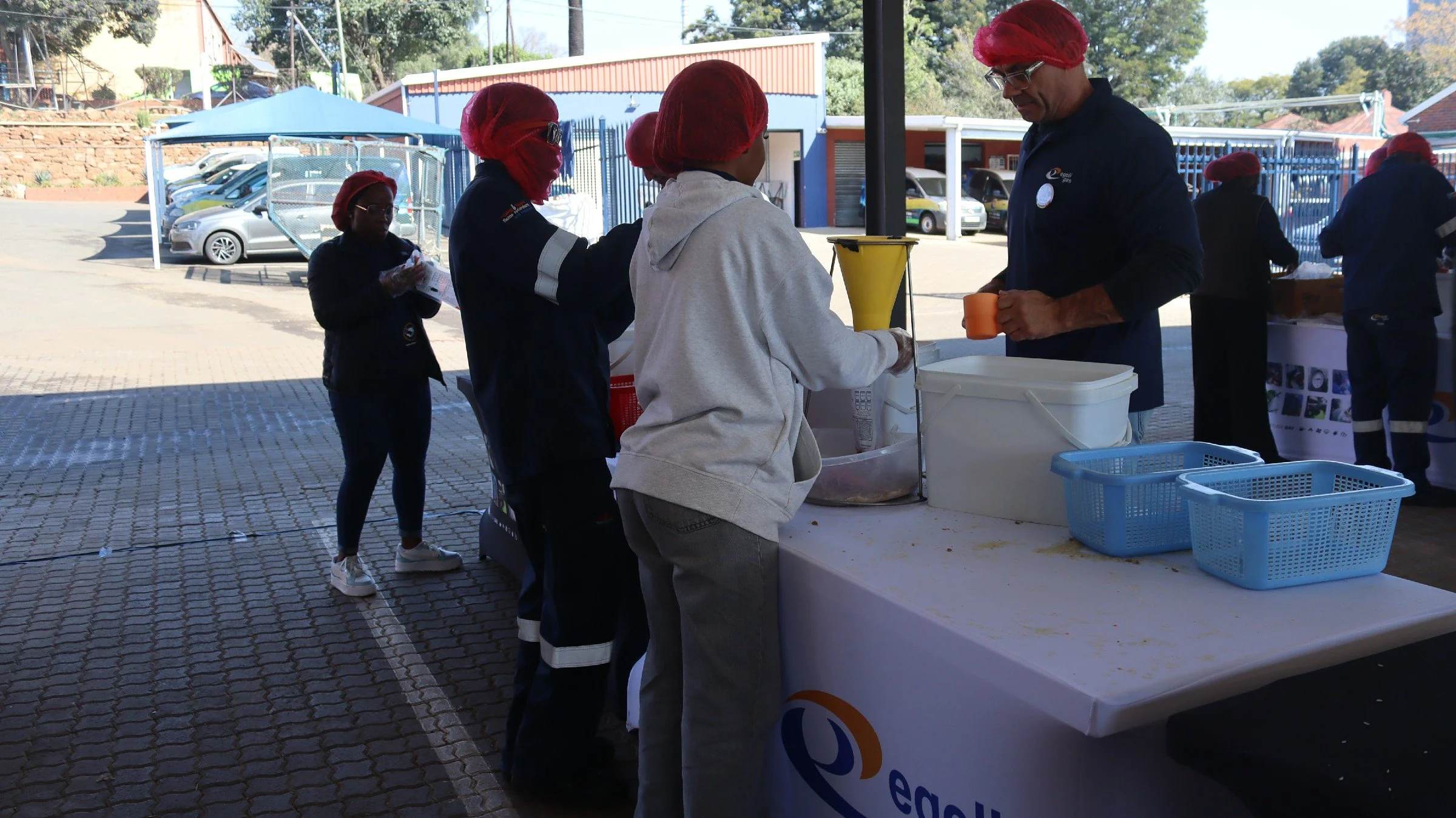 People in line at a food distribution booth outdoors, with some wearing red hair nets and uniforms, and others in casual clothing, receiving food items from a man at the table.