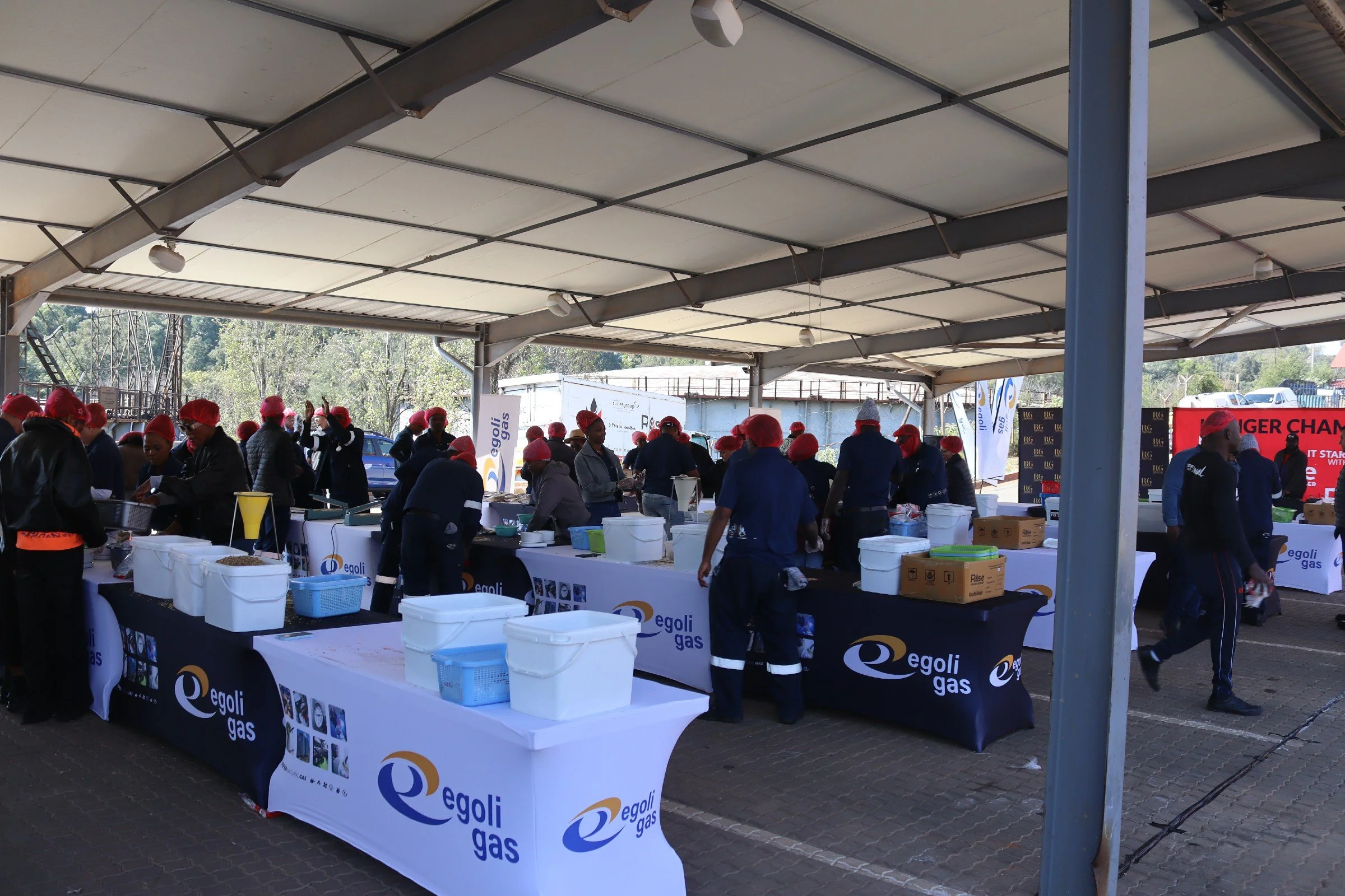 A large outdoor event with multiple tables covered with Egoli Gas banners, surrounded by many people wearing red hats, some working behind the tables and others browsing or walking around. The event is under a large canopy with trees and vehicles vis