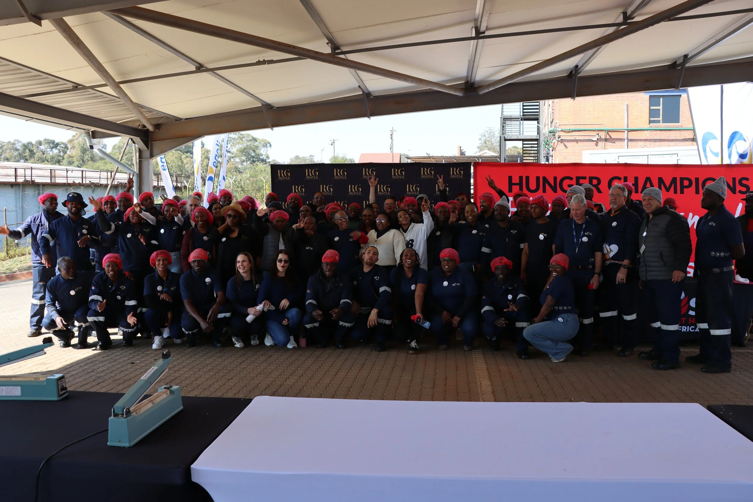 Group of people posing under a large tent with a 'Hunger Champions' banner in the background, some wearing pink hair caps and matching uniforms, in an outdoor setting.