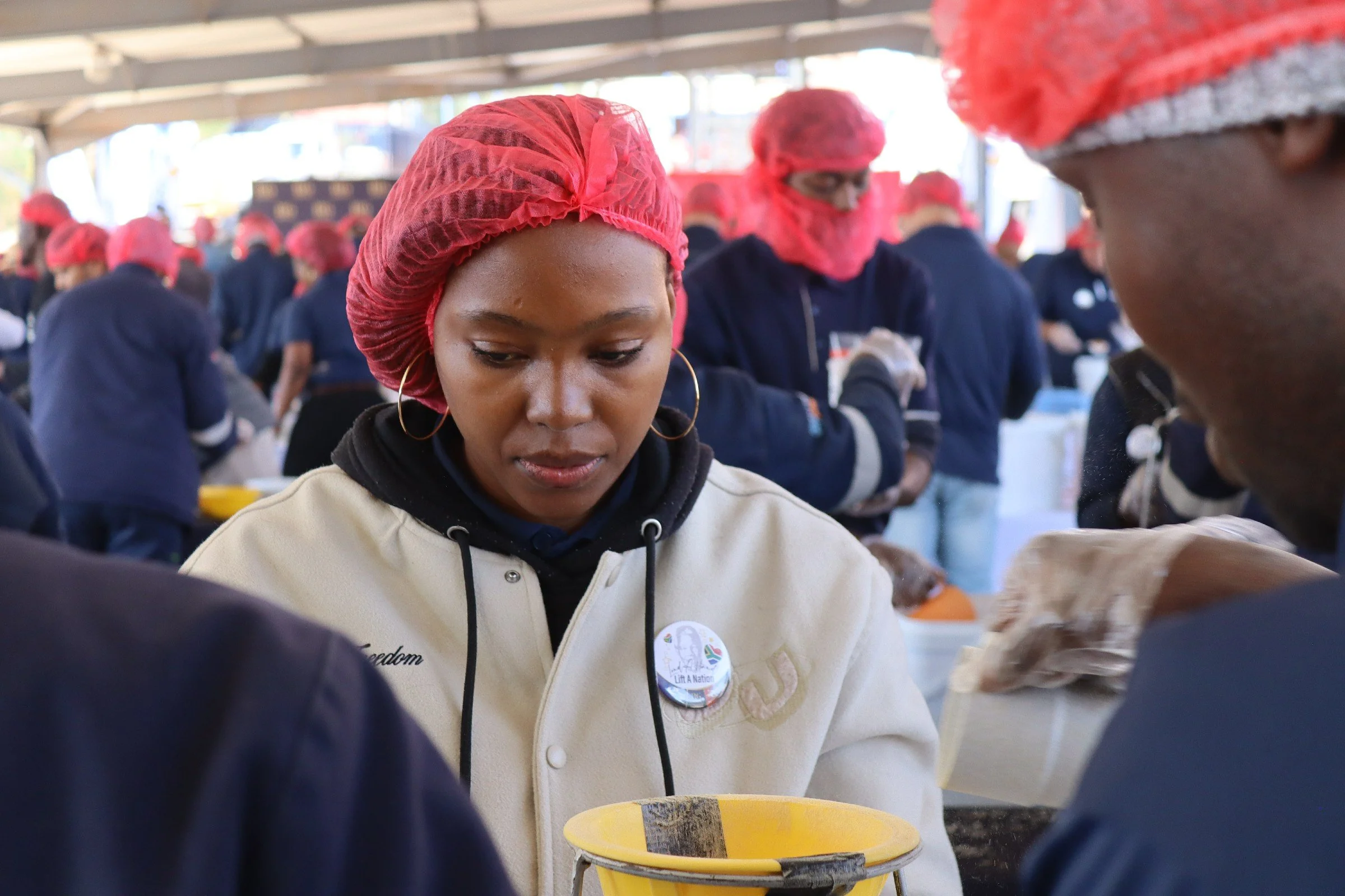 A group of people wearing red hairnets gather around a table, focusing on a task. The woman in the foreground has hoop earrings and a beige jacket with a badge that says 'Freedom.'