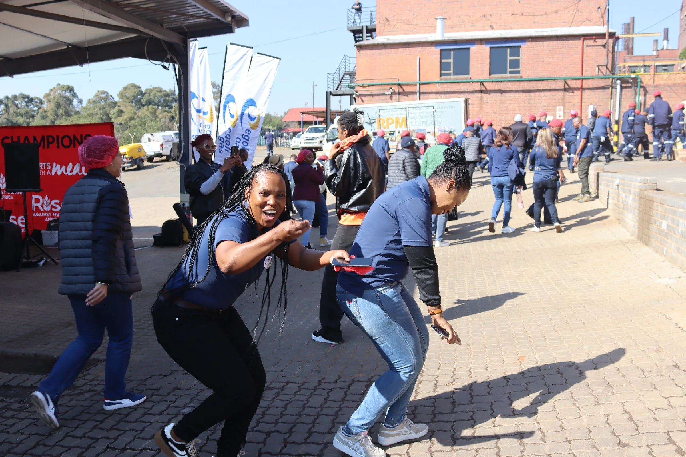 People gather outdoors near a brick building, some walking and others taking photos, with a woman in the foreground smiling and looking at the camera, wearing a dark top and braided hair.