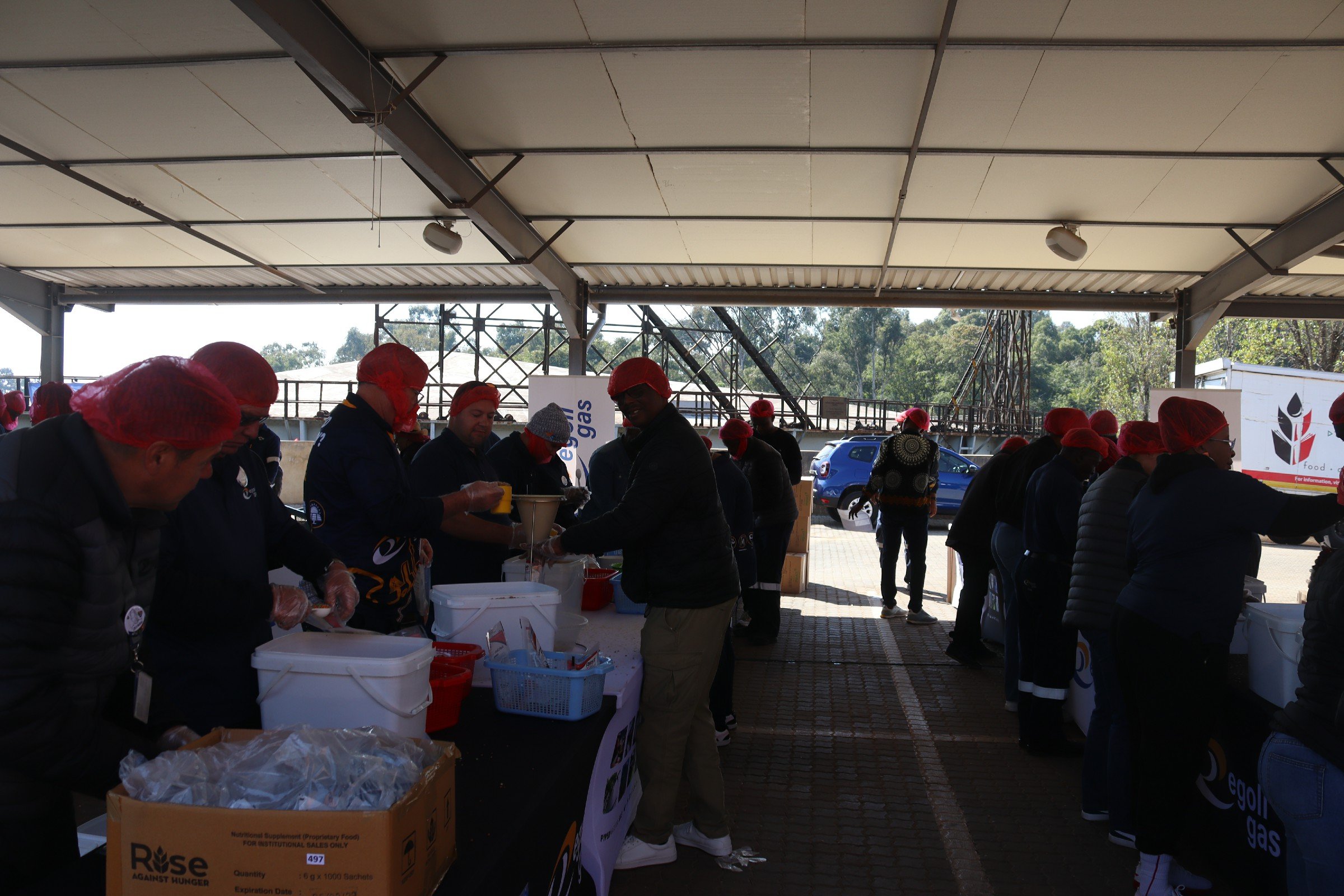 A group of people wearing red hairnets gathered under a large covered outdoor area, working with boxes of supplies, with trees and a few cars visible in the background.