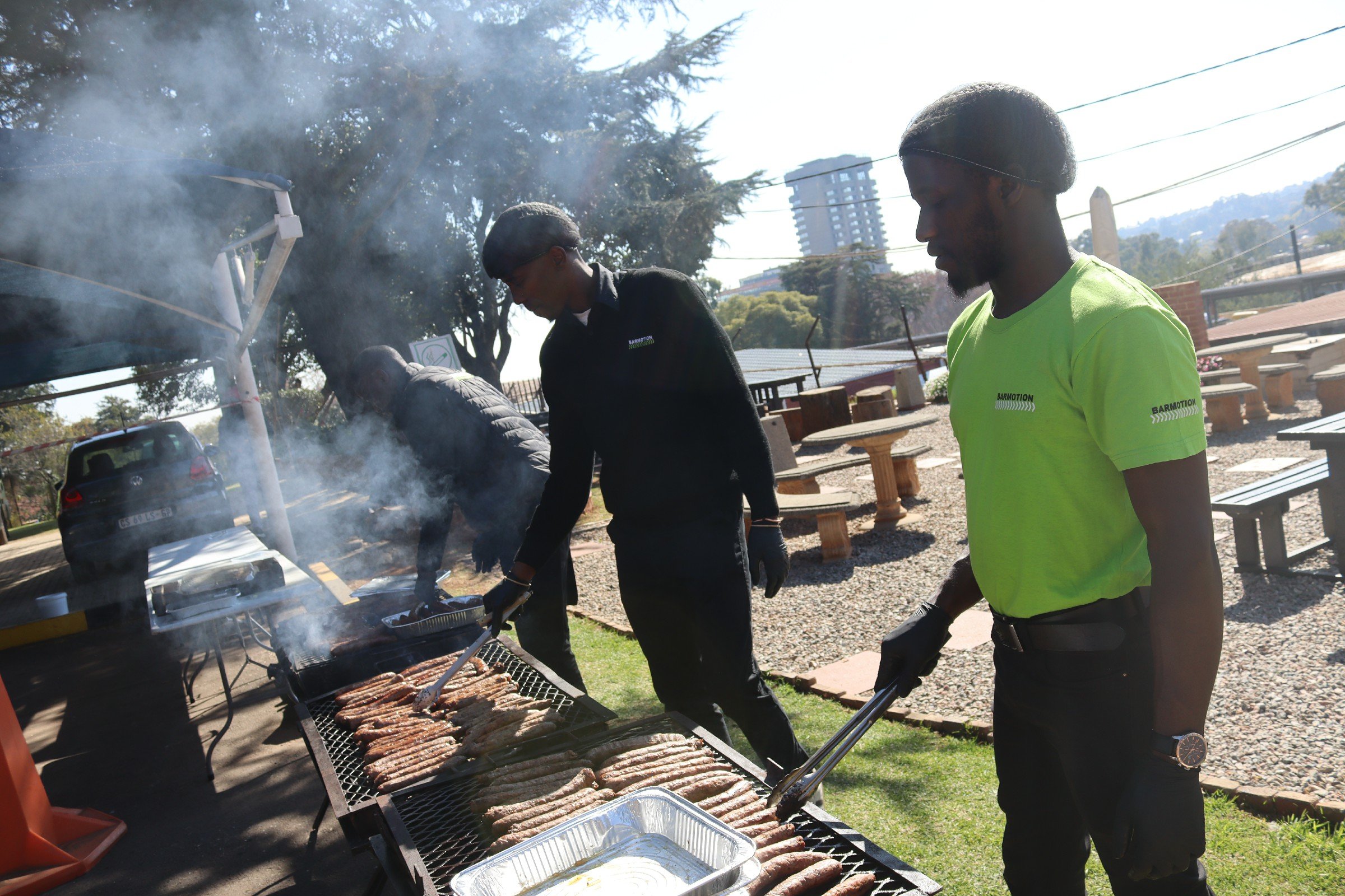 People grilling sausages and meat outdoors at a barbecue event with trees and a cityscape in the background.