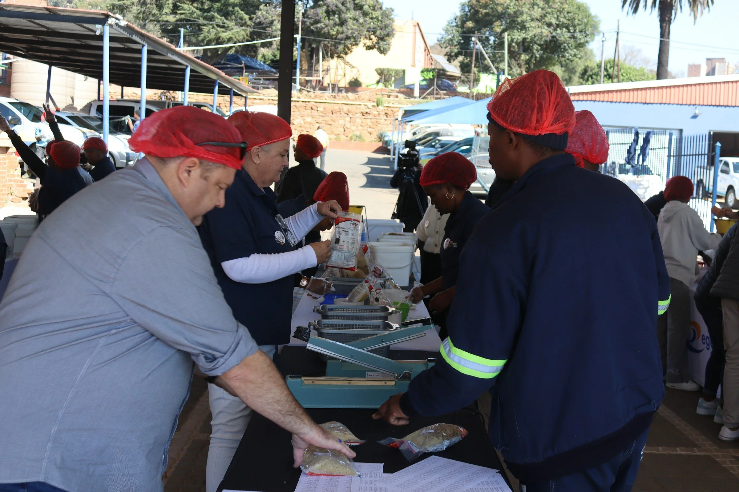 People wearing red hair nets and gloves serving food at an outdoor event under a shaded area with parked cars and trees in the background.