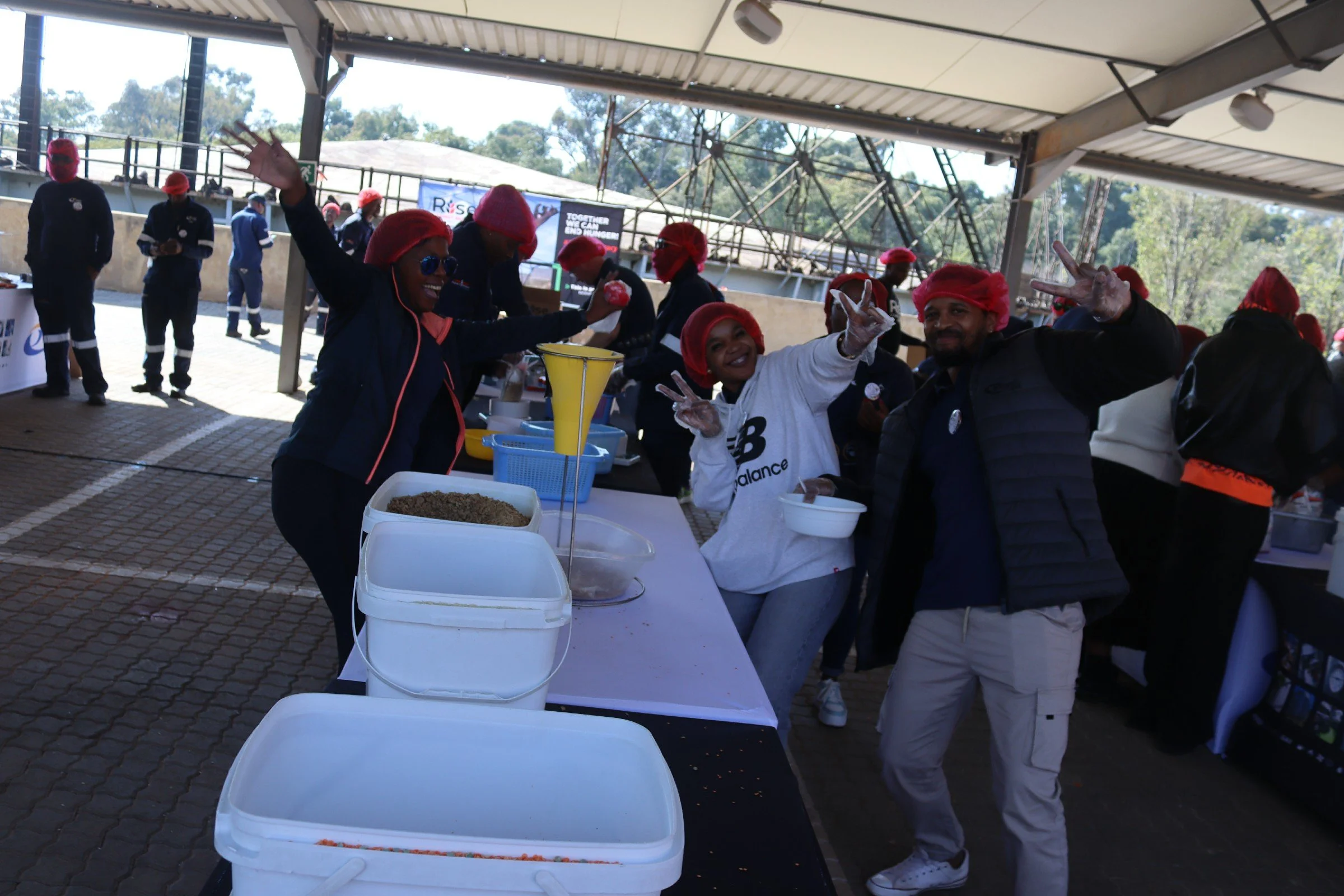People under a sheltered outdoor area wearing red hair coverings and dark clothing, some making peace signs, standing behind a table with various containers, with trees and a structure visible in the background.