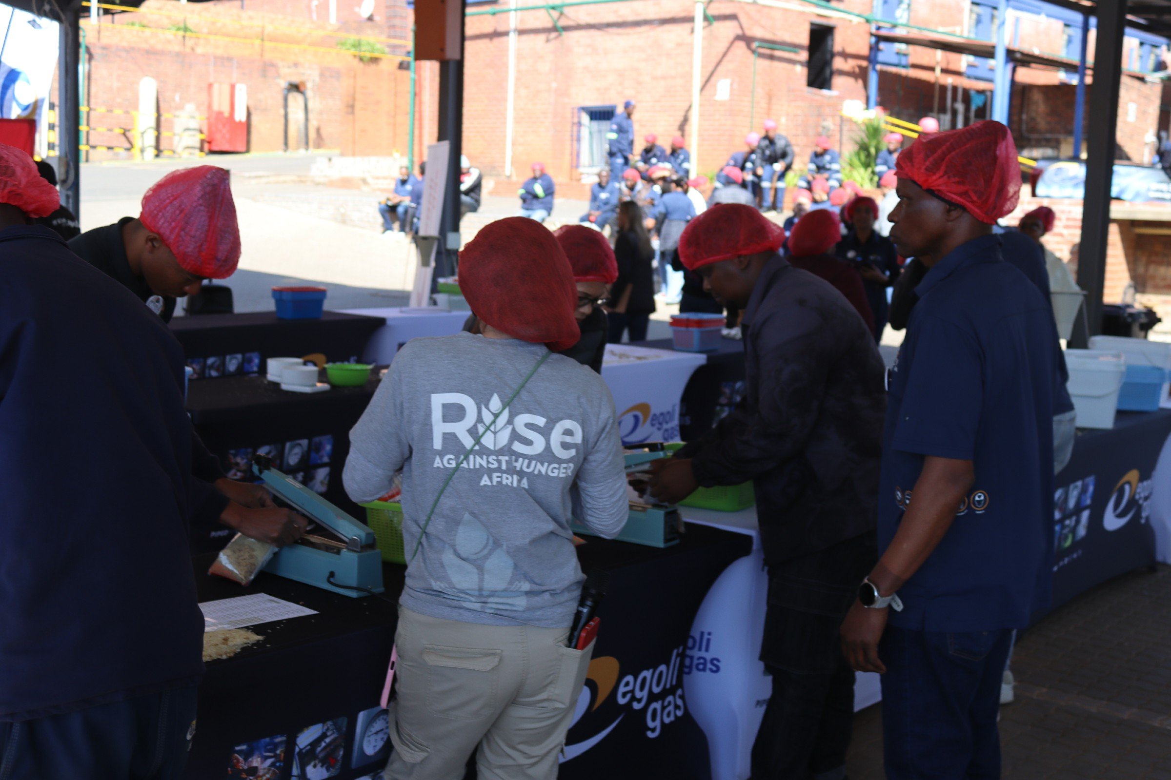 People wearing red hair nets gathered at a table during an outdoor community event. In the background, more people with red hair nets are seen sitting and standing near a brick building.