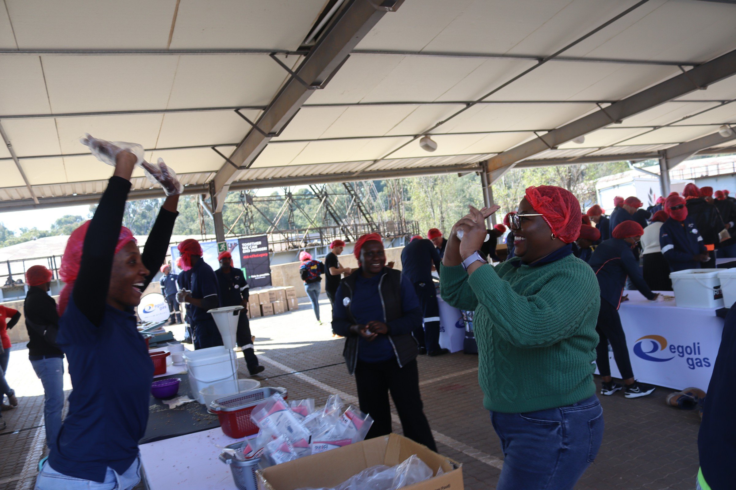 Group of people wearing red hairnets and masks working at an outdoor event under a large pavilion, with tables, boxes, and supplies around them.