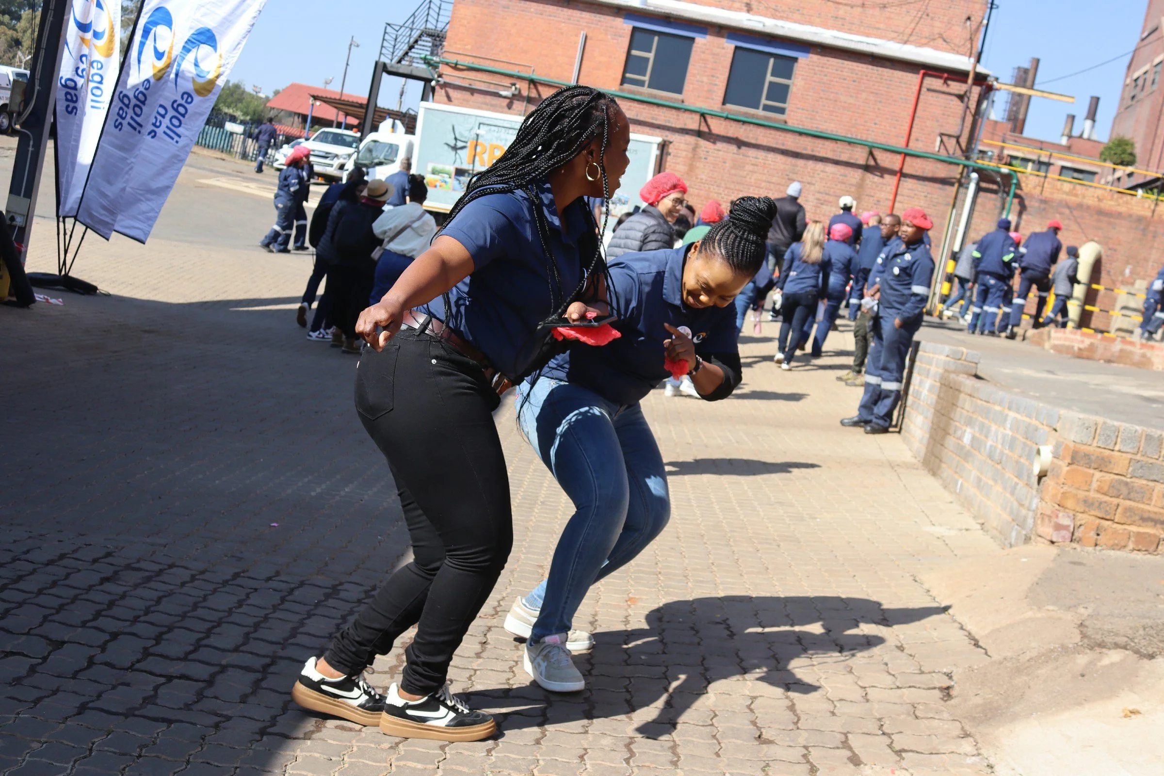 Two women dancing outdoors on a paved area during a public event, with a group of people in uniforms and red hats in the background.