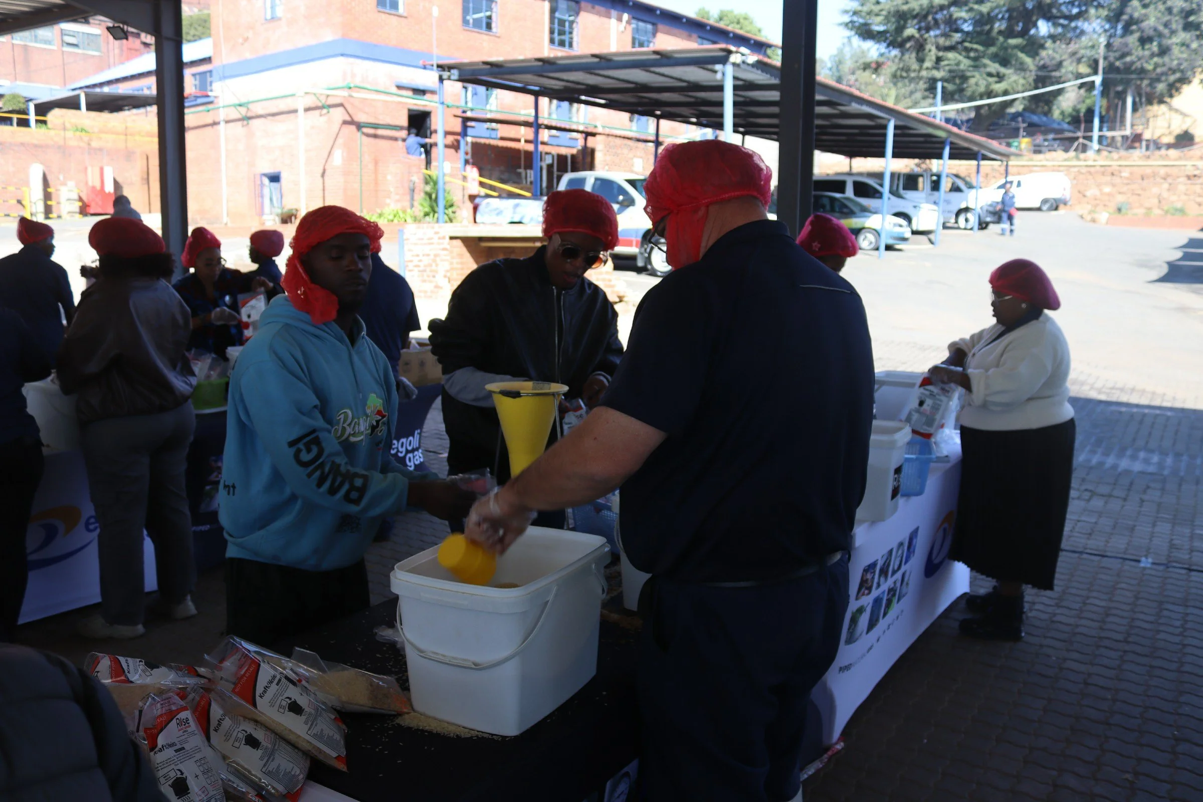 People wearing red hairnets and dark clothing gather at an outdoor table, filling plastic bags with food items, under a covered area with a parking lot and brick buildings in the background.