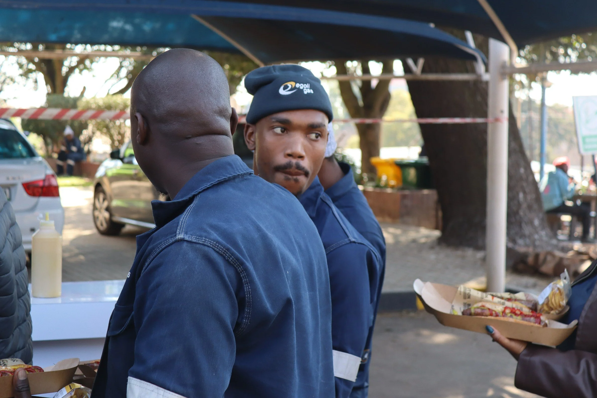 Two men facing each other outdoors, one in profile and the other looking toward the camera, with trees and cars in the background. One man is wearing a dark beanie, and the other has a shaved head.