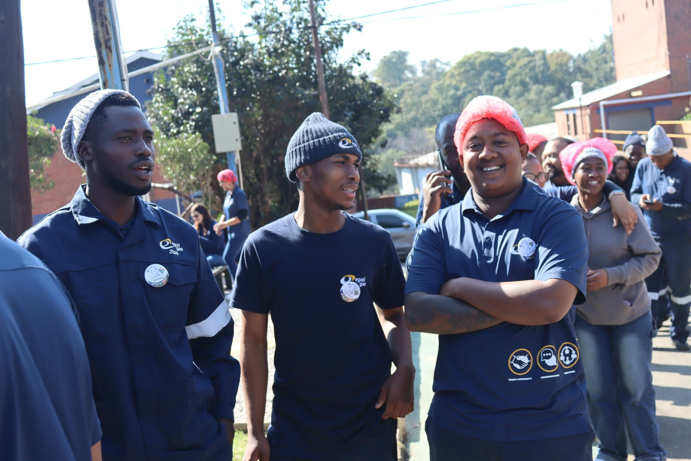 Group of people outdoors, some wearing matching navy blue shirts with logos, during daytime. One man, wearing a pink shower cap, smiling with folded arms. Others wearing beanies, some using phones, in front of trees and buildings.