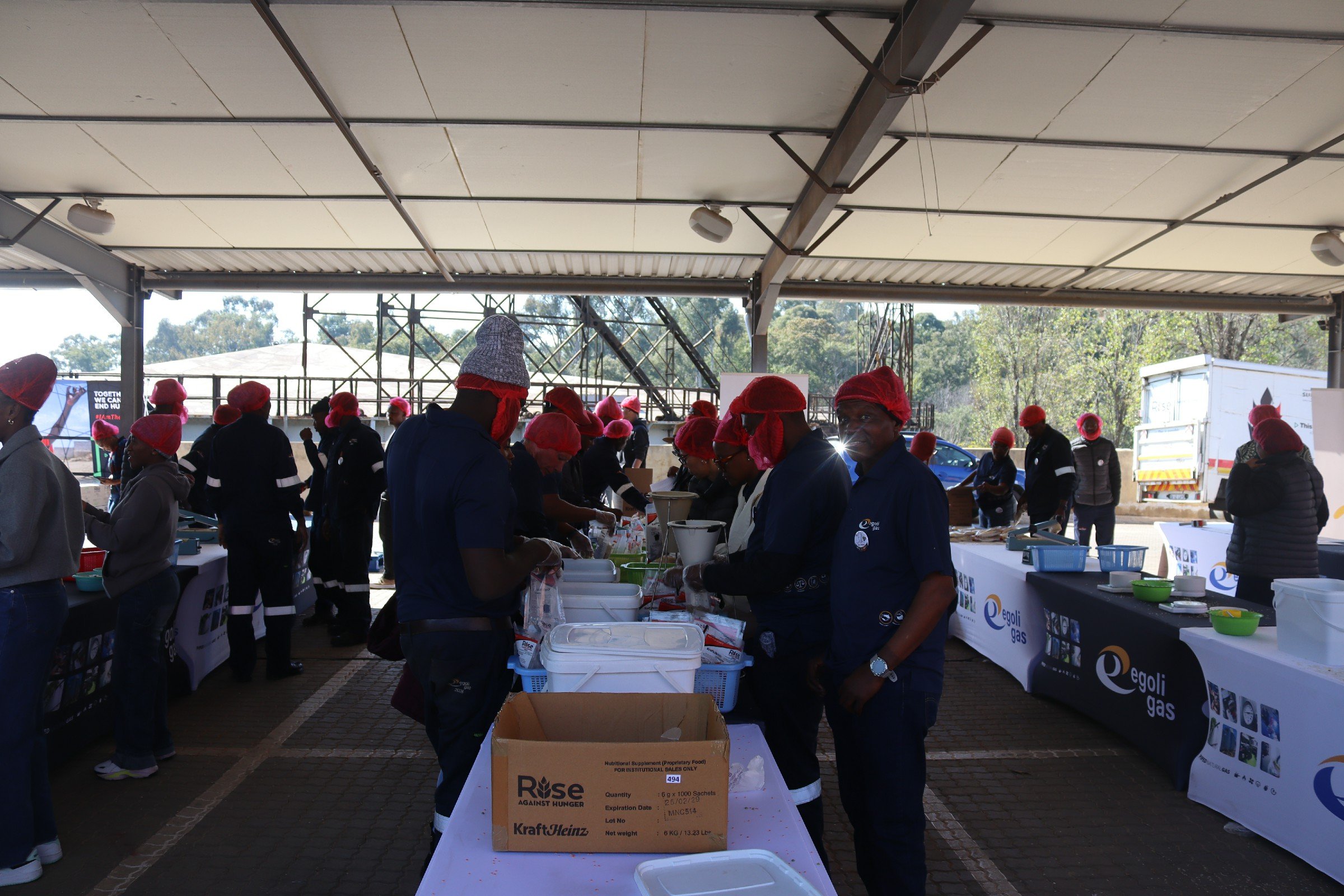 People wearing red hair coverings and dark uniforms at a food distribution event under a large shelter, with tables displaying food items and banners with logos, and trees in the background.
